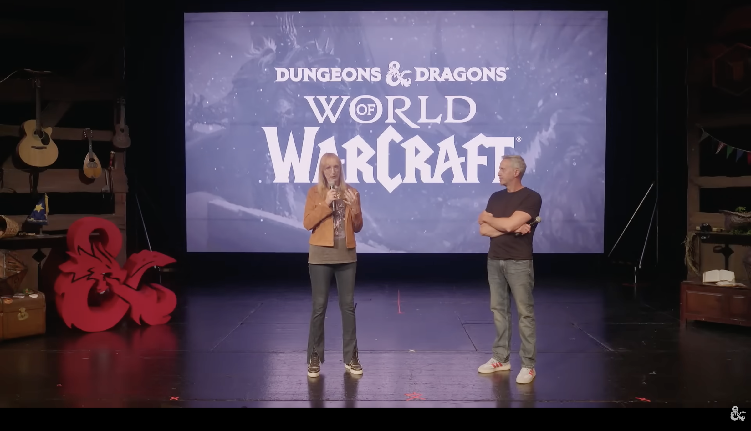 Two people stand on a stage decorated with the Dungeon & Dragon ampersand in front of a screen that reads "Dungeons & Dragons: World of Warcraft".