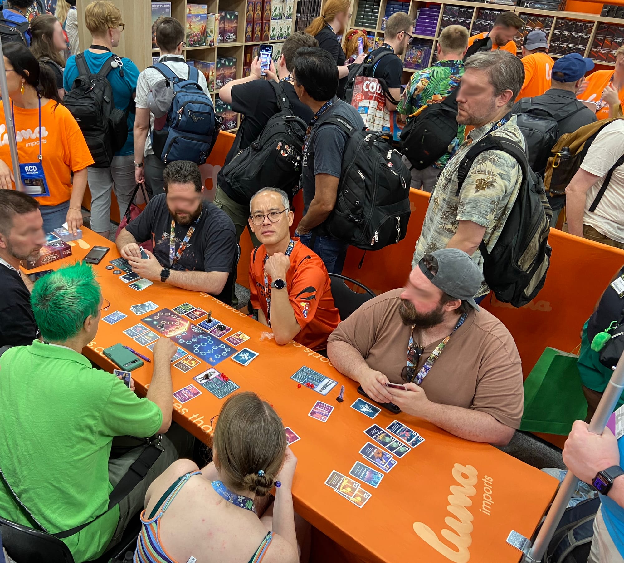 People sit around a table playing the 2026 version of Not Alone in the Luma Imports booth at Gen Con 2026, with a line of people weaving through the tables. One man stares at the camera with a "Whyyyyy?!" expression on his face.