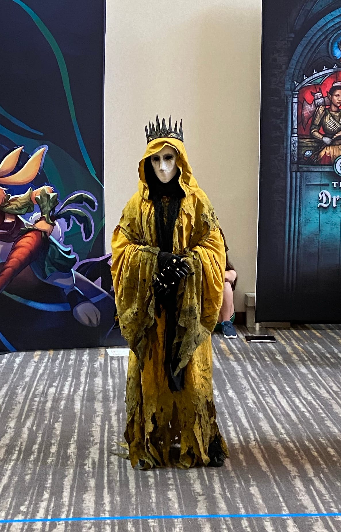 A figure in a stained and torn yellow cloak stands with a pointy crown on their head and their clawed hands holding one another. A white mask hides their face, revealing only shadowed eyes.