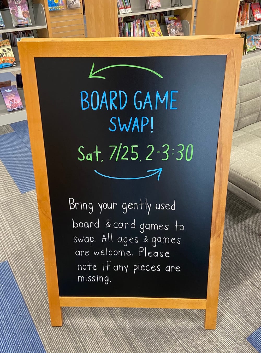 A sign reads "Board game swap! Sat. July 25, 2-3:30. Bring your gently used board & card games to swap. All ages & games are welcome. Please note if any pieces are missing."