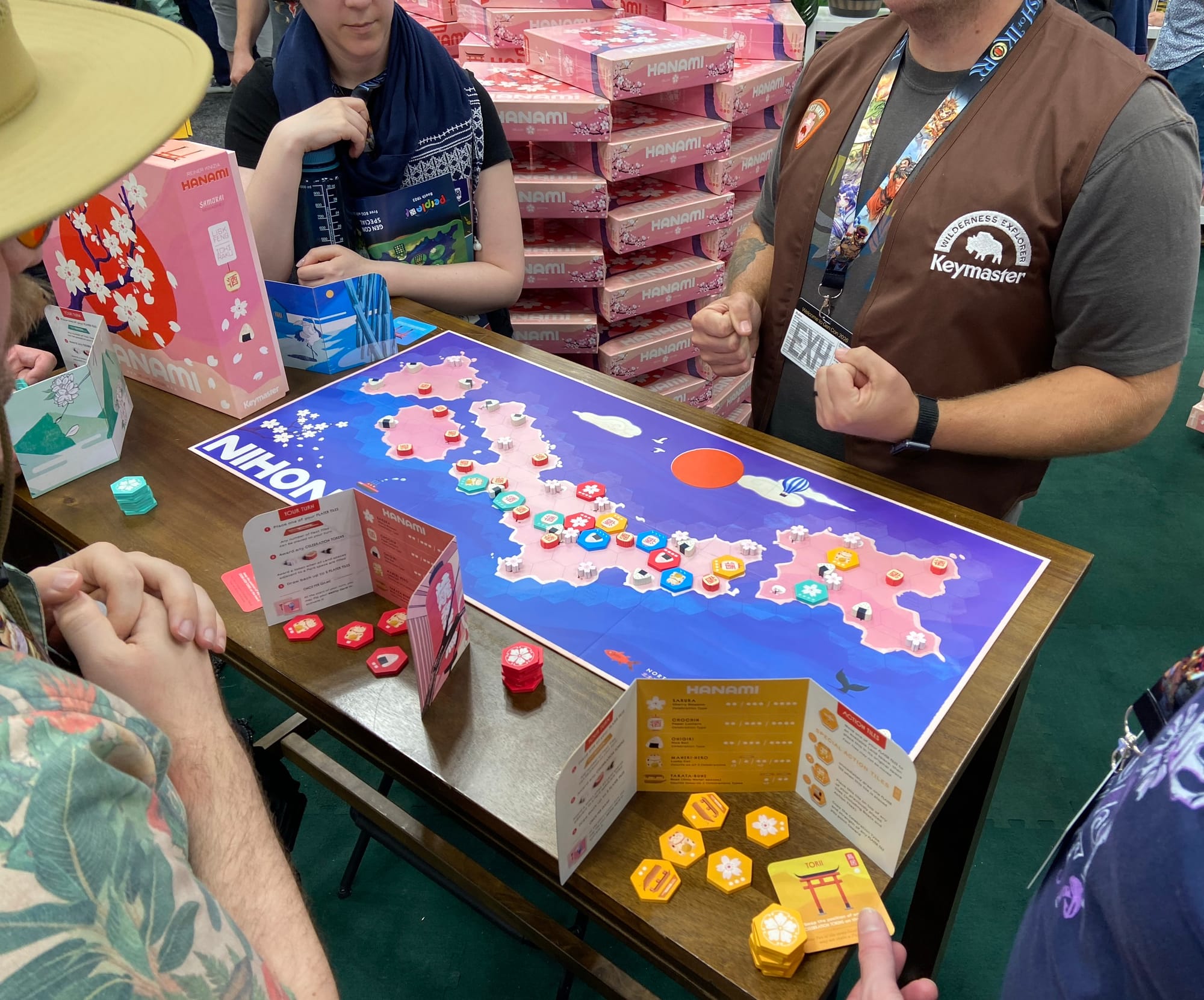 The game board for Hanami fills the table, with player tokens behind screens around the game board and people standing around the table, with a giant stack of Hanami in the background.