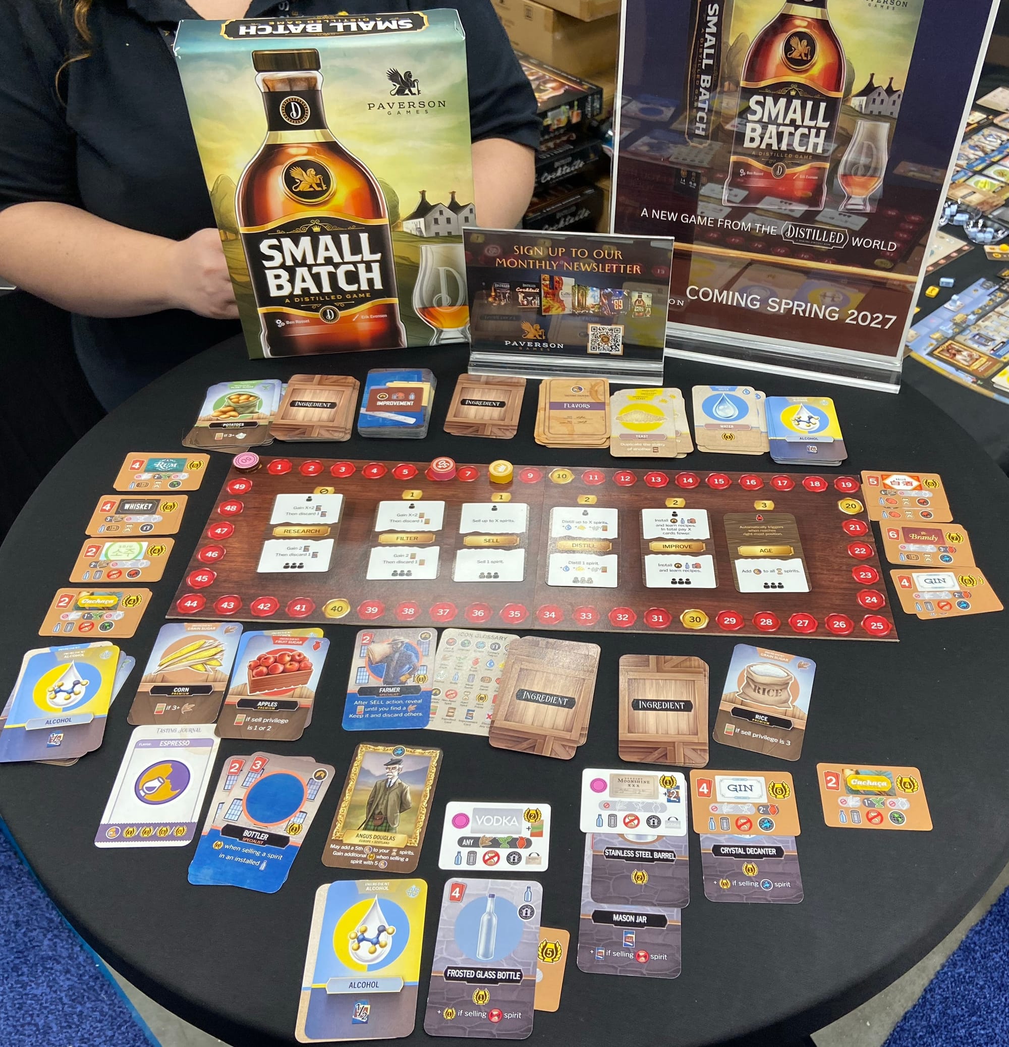 The game box for Small Batch shows a liquor bottle bearing that name. On the table in front of the box is a game board and dozens of cards.