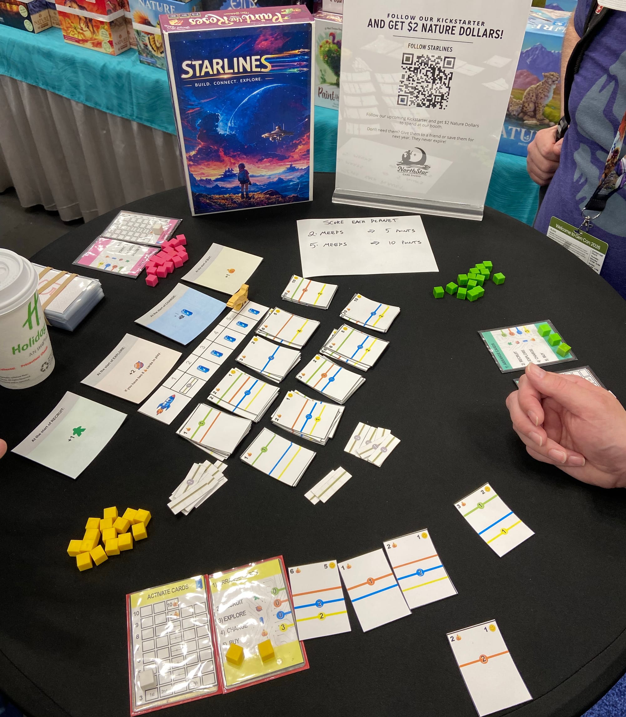 Dozens of pieces of paper are stacked on a table, with each piece having 1-4 colored lines on it. One person's play area shows cards placed in a row, creating continuous colored lines across the cards.