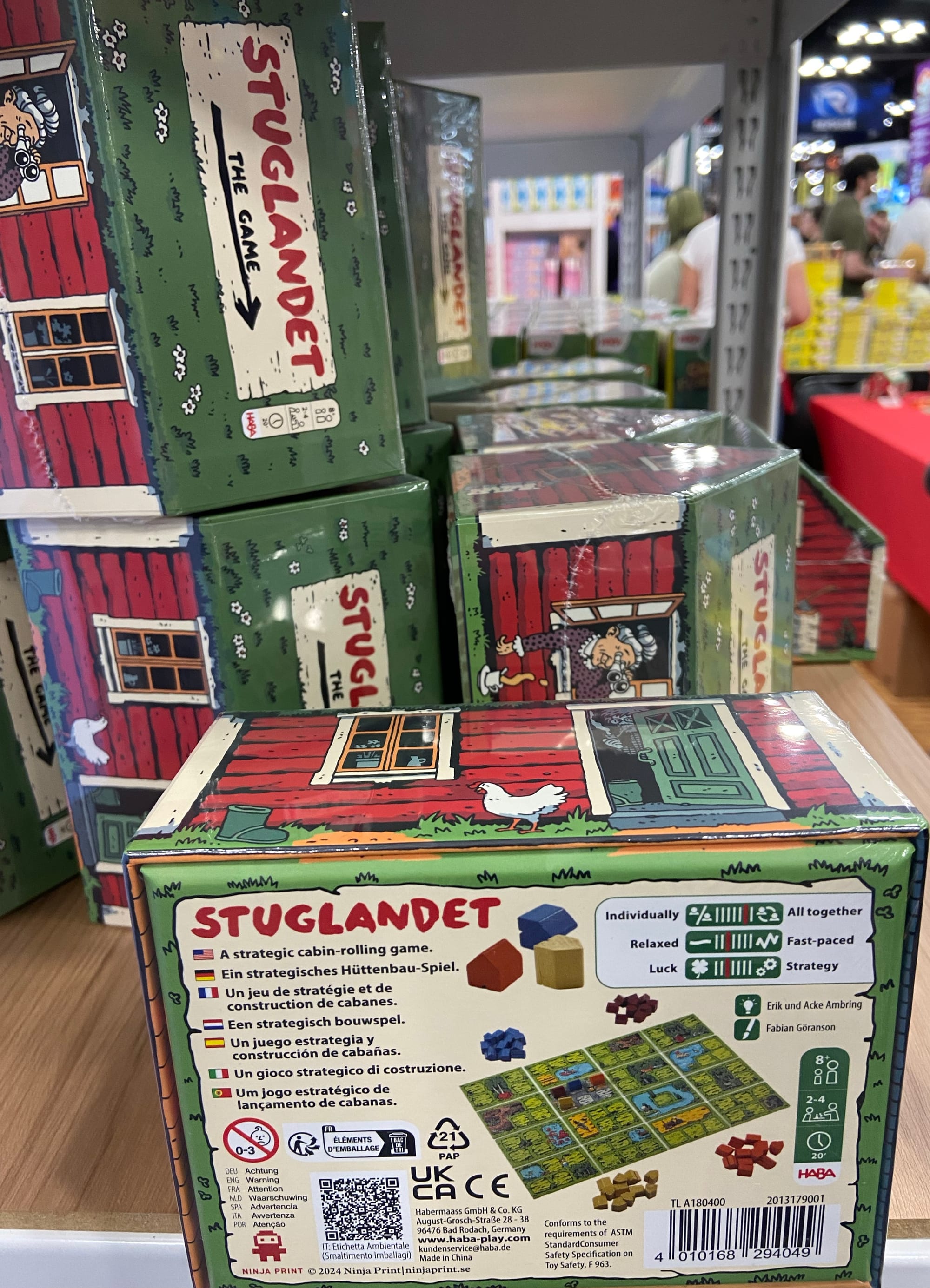 Multiple boxes of the game Stuglandet, which are shaped like a log cabin house with a slanted roof, are stacked on a shelf.