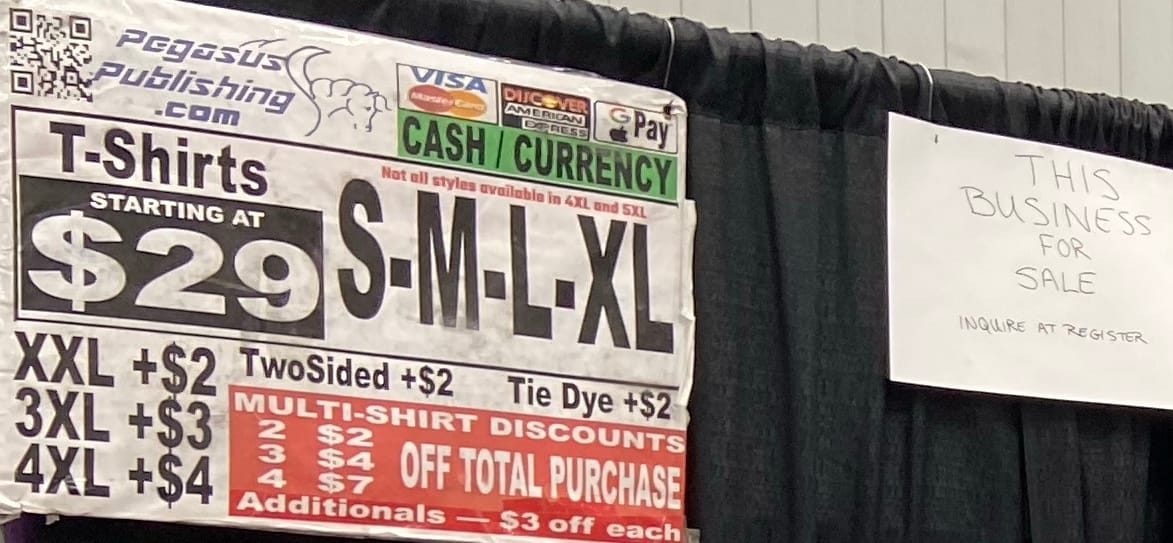 A zoomed-in image of the T-shirt booth focusing on a sign that reads "This business for sale; inquire at register".