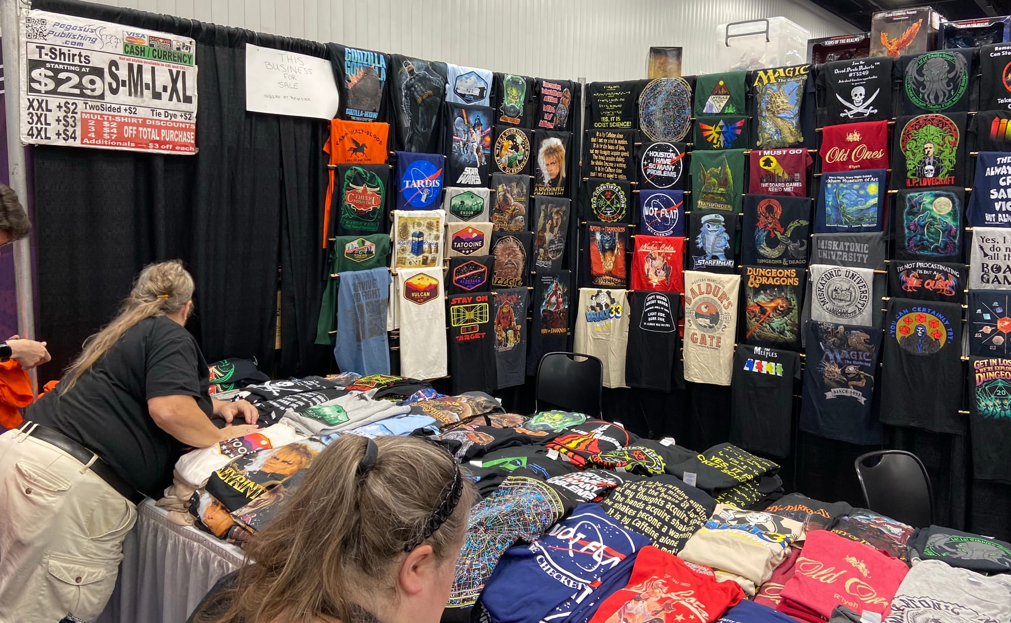 Dozens of T-shirts with fantasy images and sayings are draped over the cloth walls of a booth; many more such shirts fill the tables in that booth.