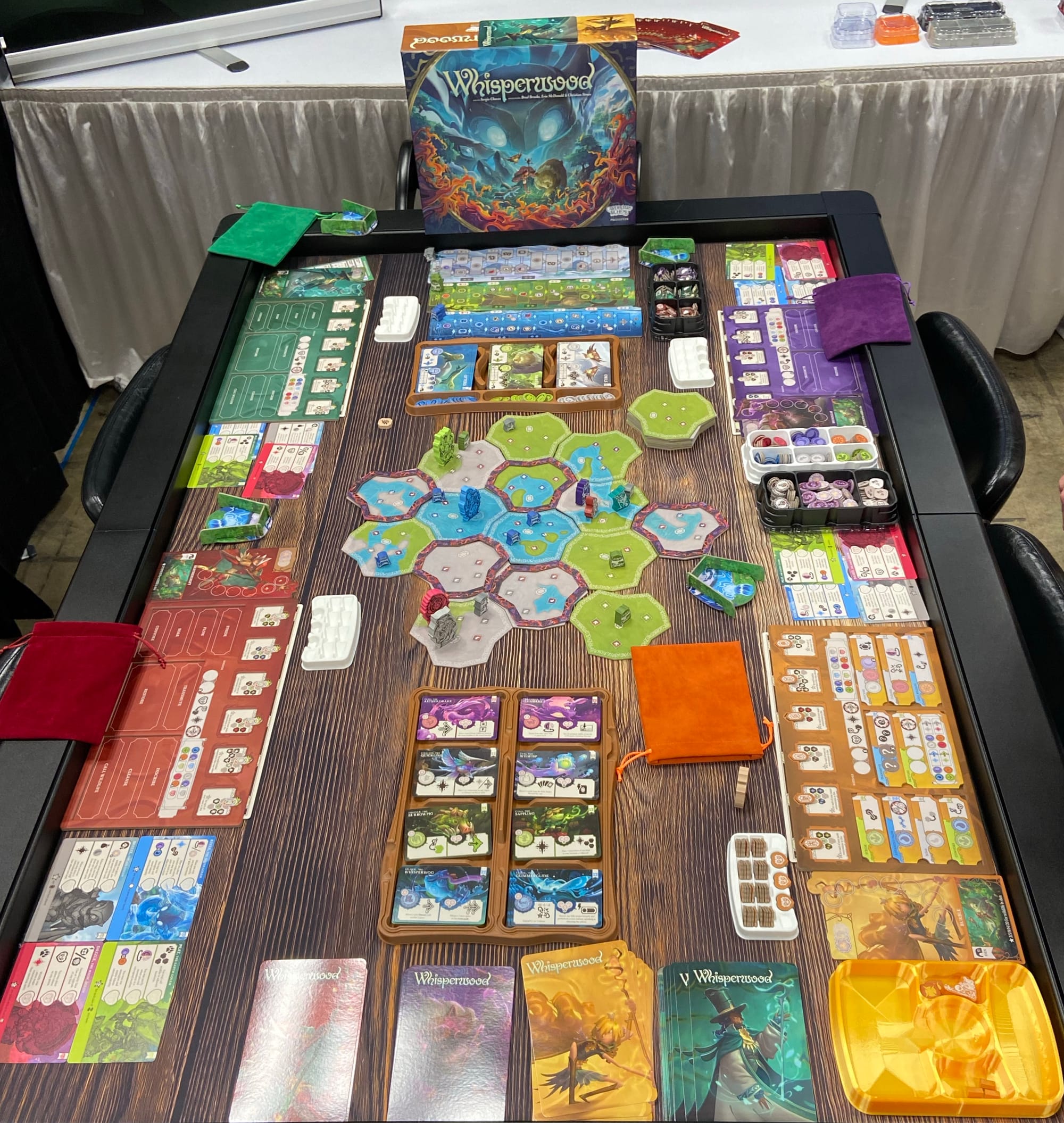 A six-foot table has nearly every surface covered with components of Whisperwood, including a character board, mat, and other cards for each player, multiple card trays, and a playing area composed of hexagonal-ish tiles.