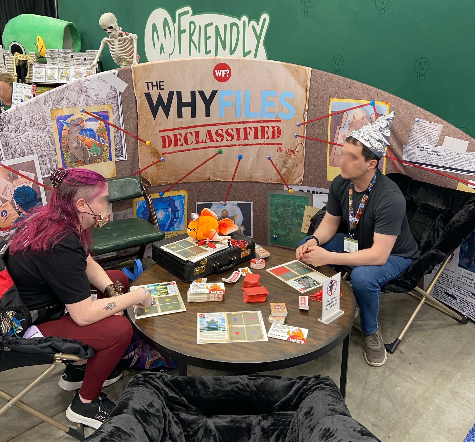 In front of a banner labeled "The Why Files Declassified", two people sit in low chairs at a table, one wearing a tinfoil hat, with cards stacked on a table between them.