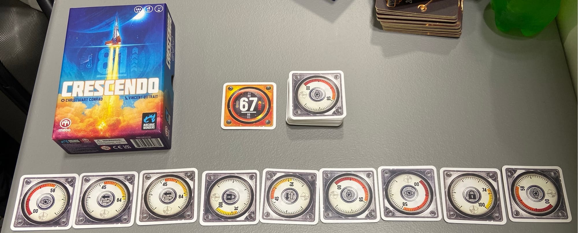 The front cover of the game Crescendo is above a row of nine cards; a deck of cards is nearby, along with a face-up card bearing the numeral 67