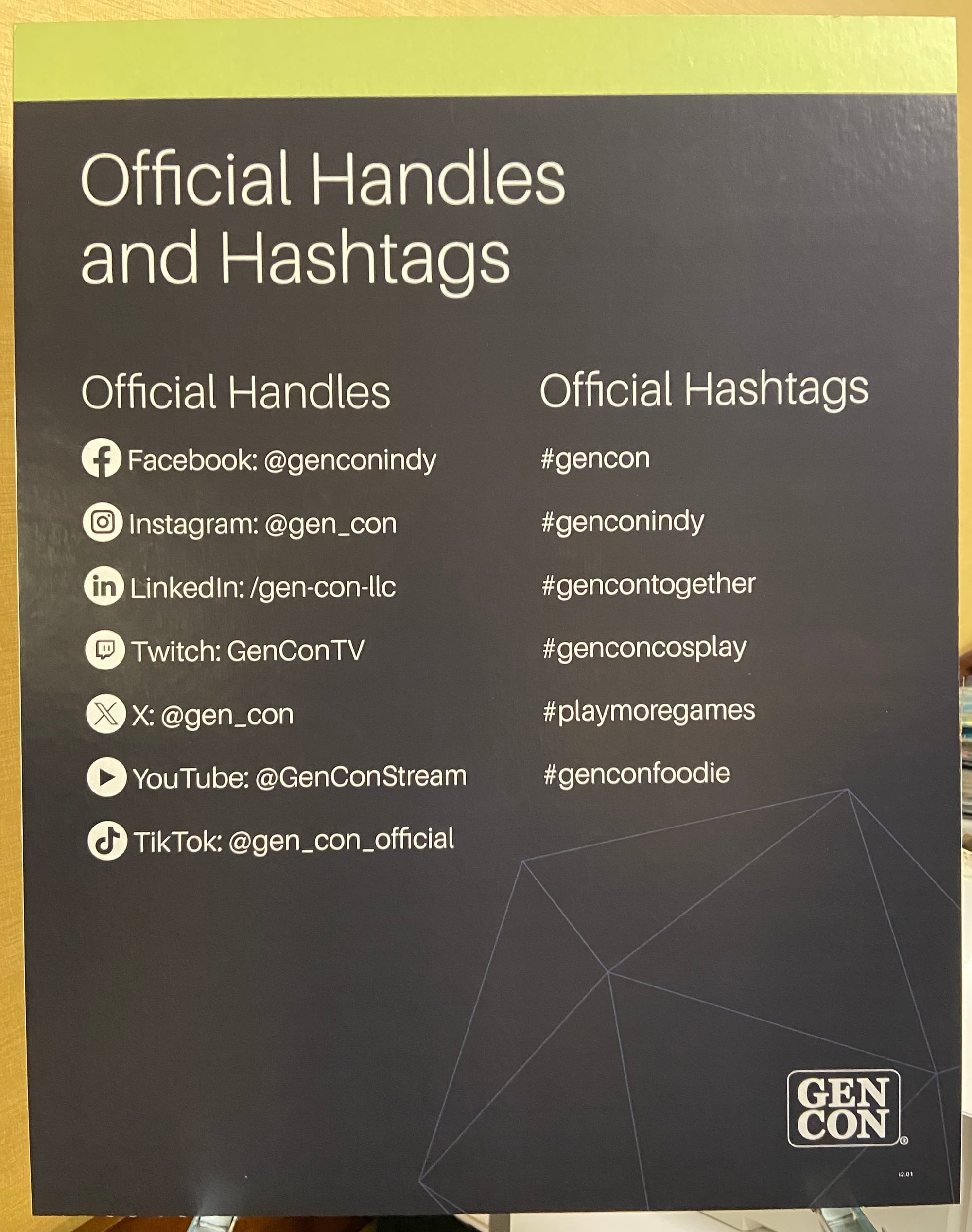 The sign reads "Official Handles and Hashtags", then lists the handles for Gen Con on Facebook, on Instagram, etc.