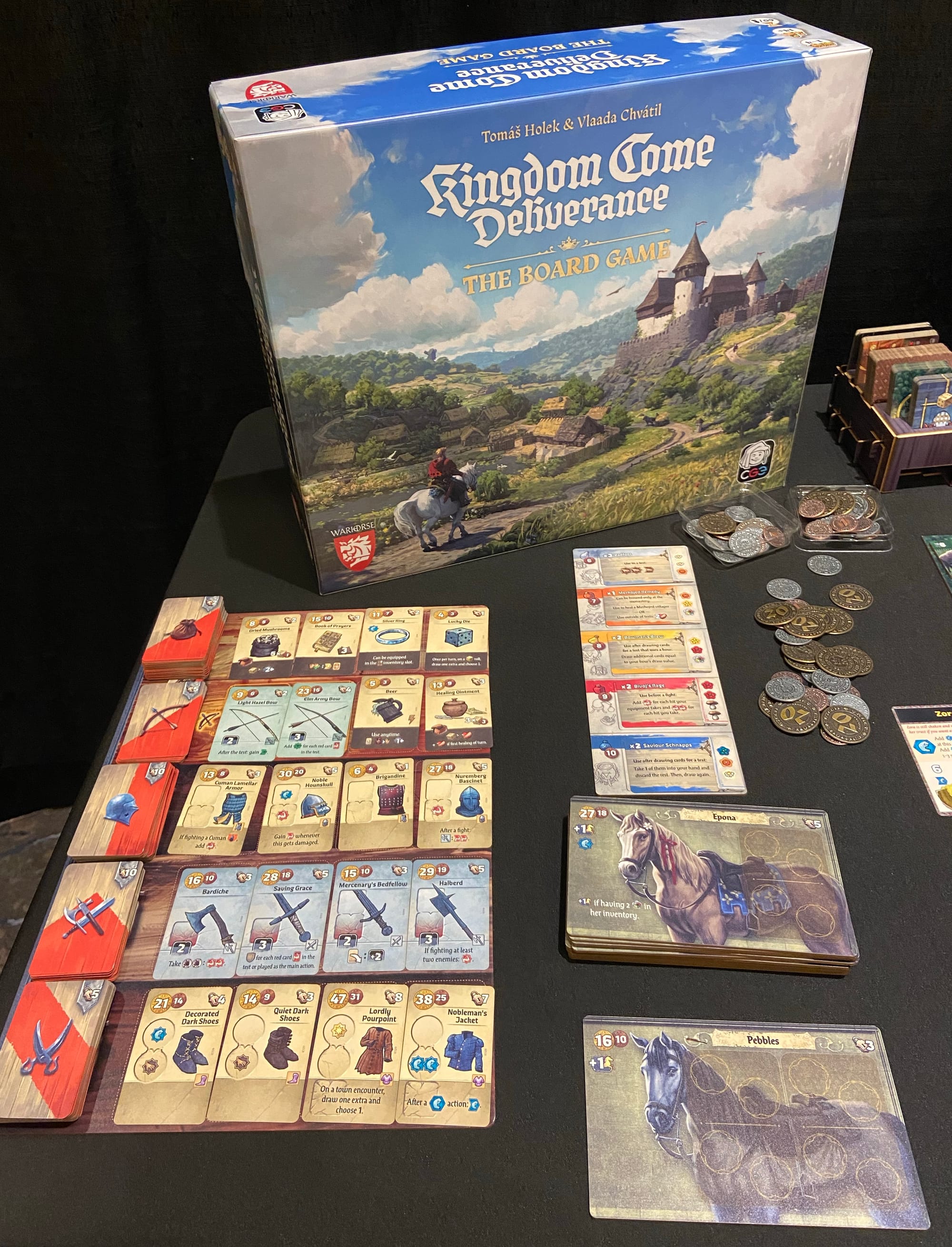 A mock-up of the Kingdom Come: Deliverance – The Board Game box, along with a market showing four items in each of five categories, a spill of metal coins, and a couple of horse tiles.
