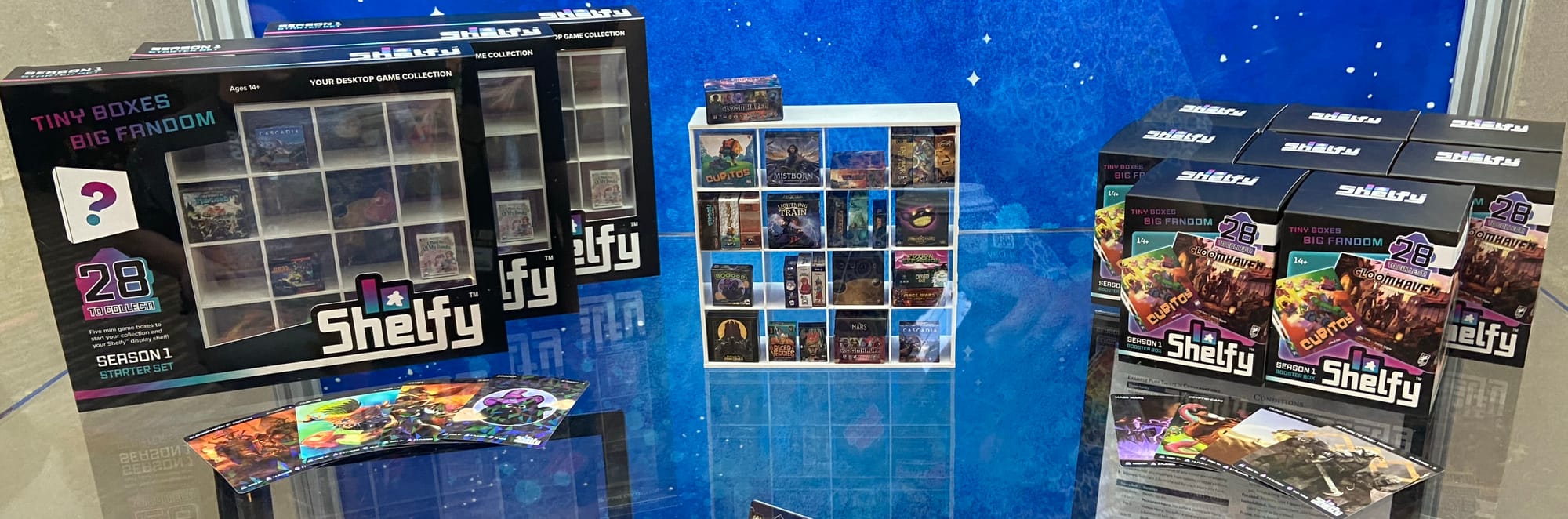 Packages for Shelfy feature a miniature Kallax-style display case with sixteen hollows for games in a 4x4 grid; Shelfy booster boxes are also depicted, along with a full display case showing all 28 boxes.