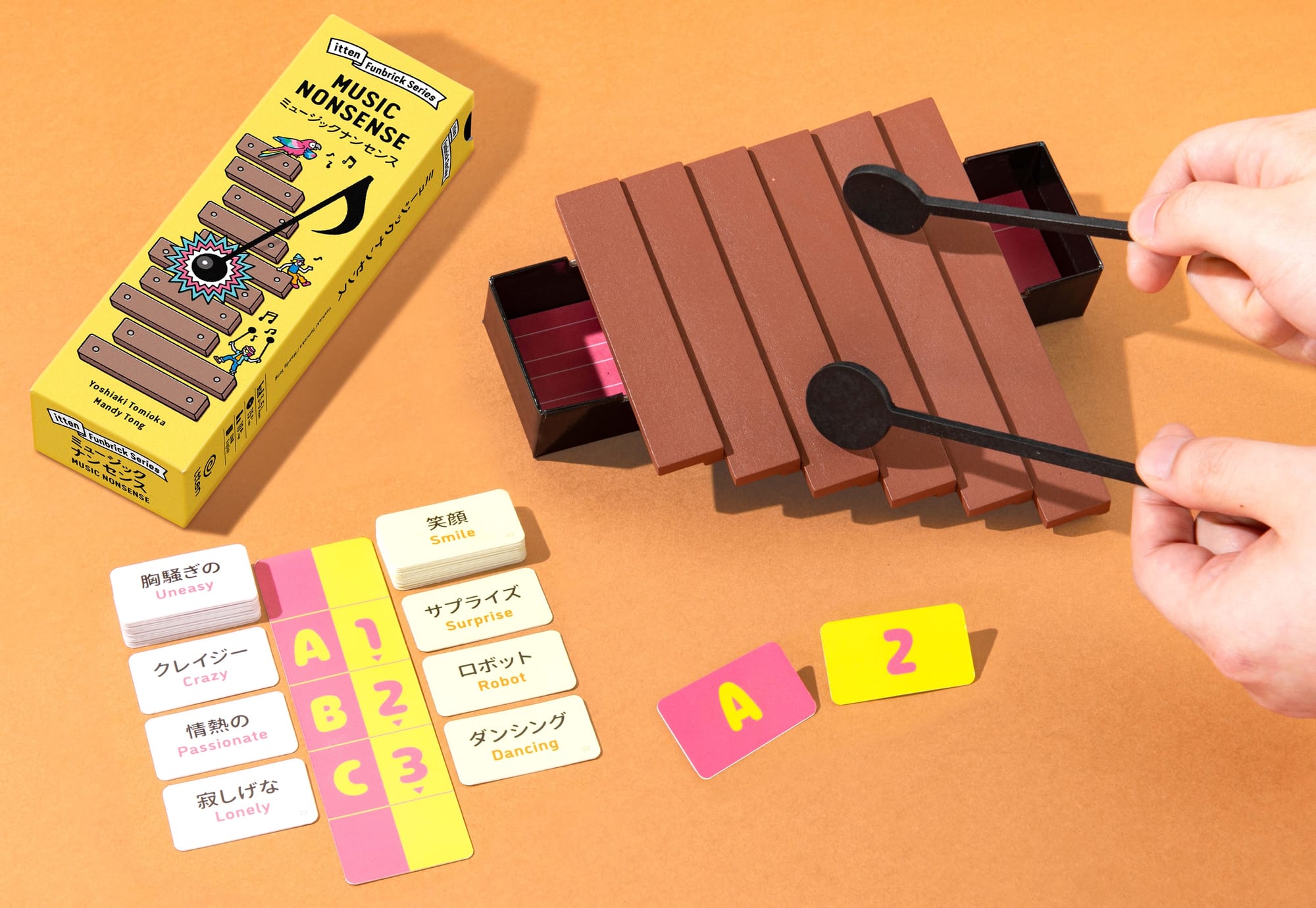 Six wooden sticks of differing lengths lay across the open box bottom, creating a xylophone that's being beat on with two wooden sticks. Two columns of cards featuring words in English and Japanese are nearby.