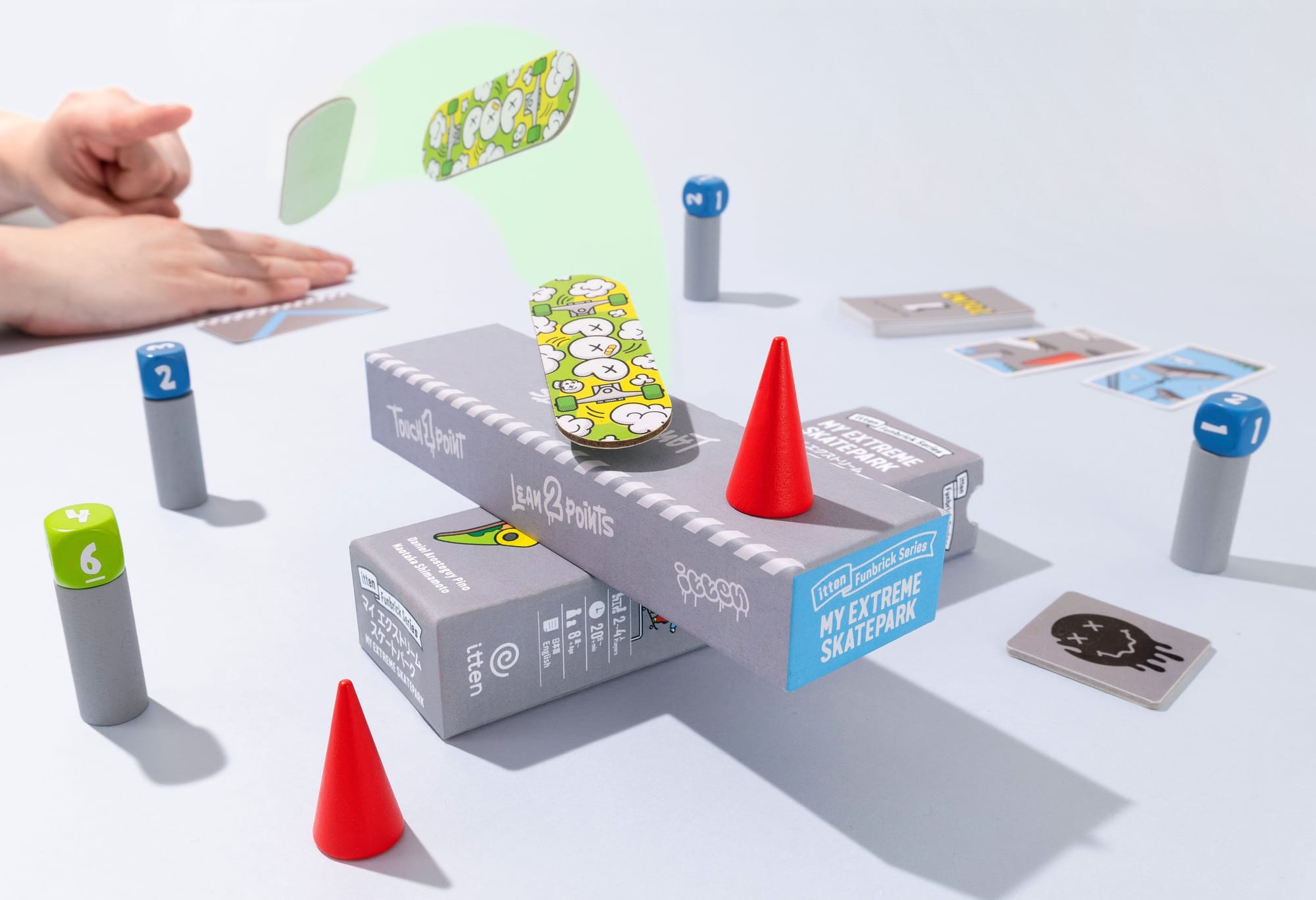 The box bottom is stacked on the box top and surrounded by wooden pillars topped with dice. A person's hands have flipped a skateboard, which flies through the air and lands on the box bottom.