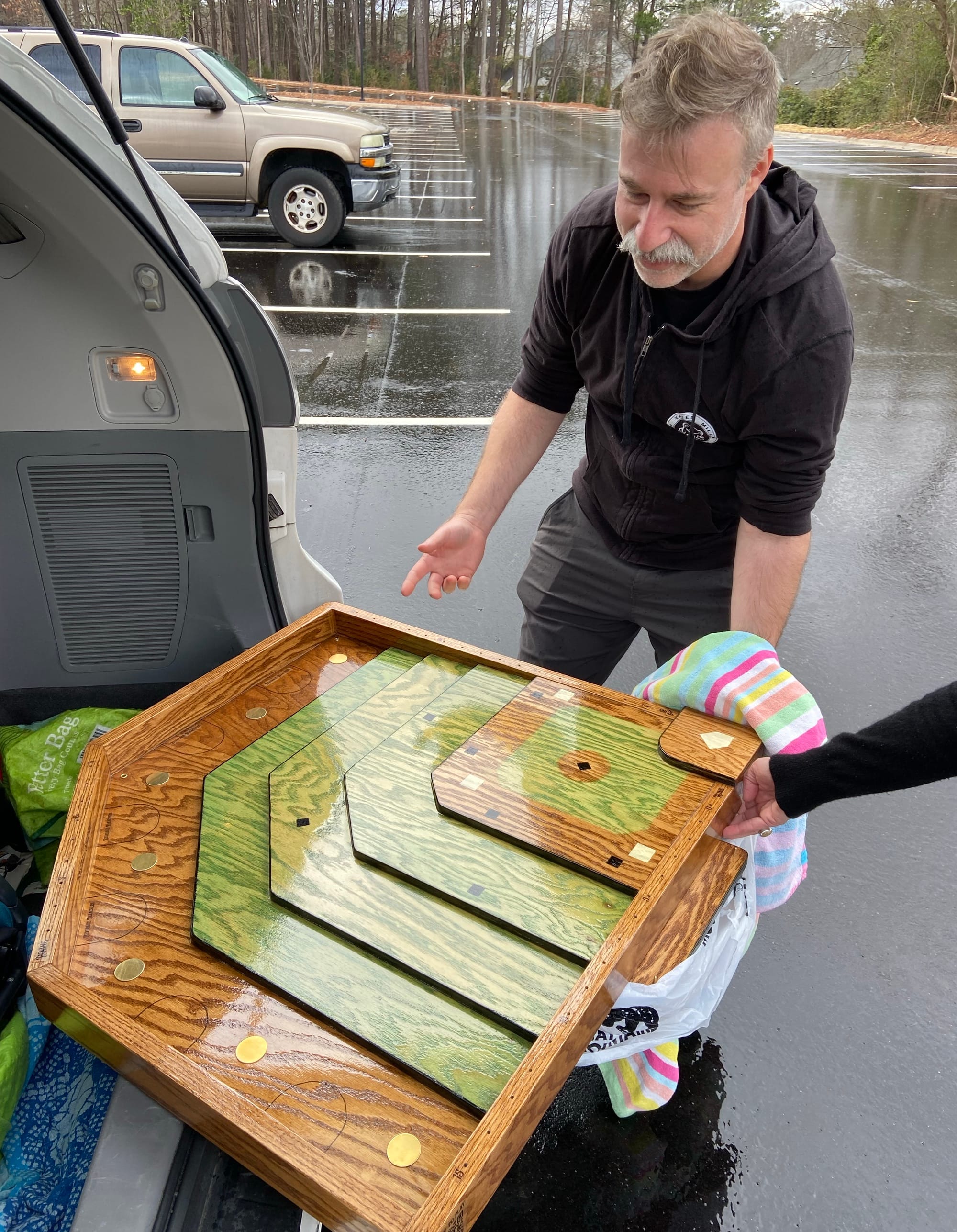 The man from the previous photo is now standing next to the back of an open SUV, holding one corner of the baseball game board while the rest is balanced on the edge of the trunk.