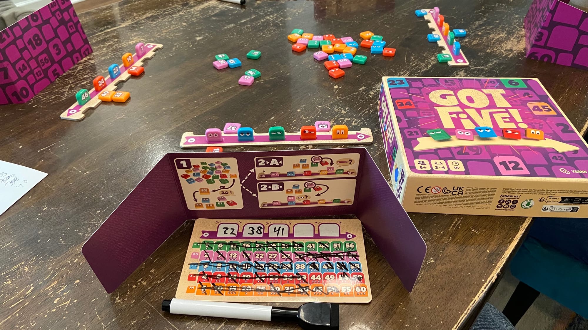 In this three-player game, each player has a rack of five numbered tiles facing away from them. You can see a deduction board behind one player's screen, with many of the numbers from 1-60 being marked out and three of the tiles being guessed.