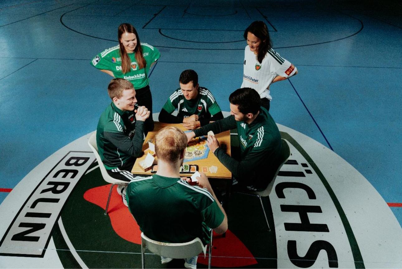 Four people in sports uniforms sit at a table on a handball court playing CATAN; two other people stand near the table watching.