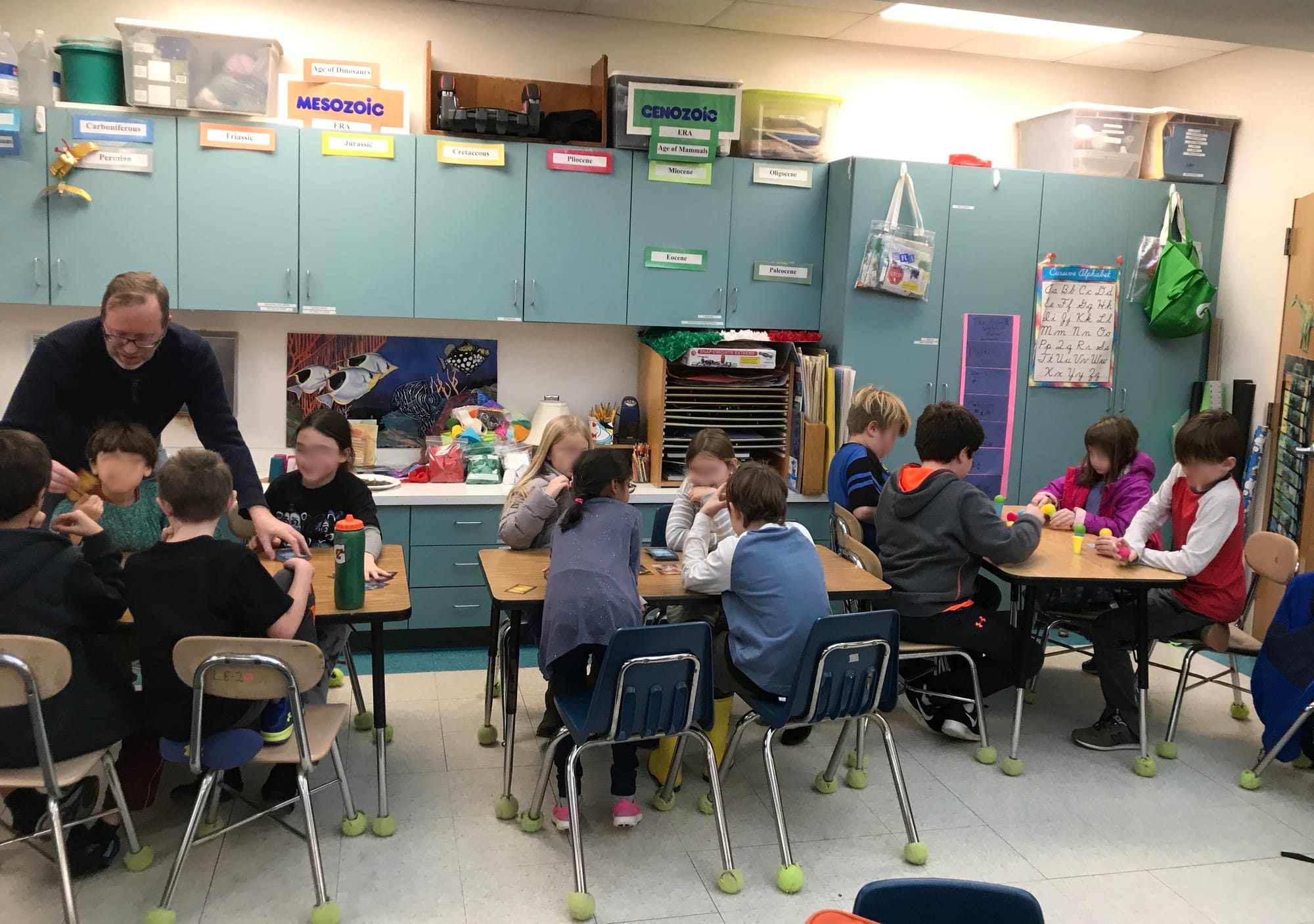 Three tables are depicted in a classroom, with four children aged 9-10 at each table; one adult stands over a table, apparently instructing kids on how to play.