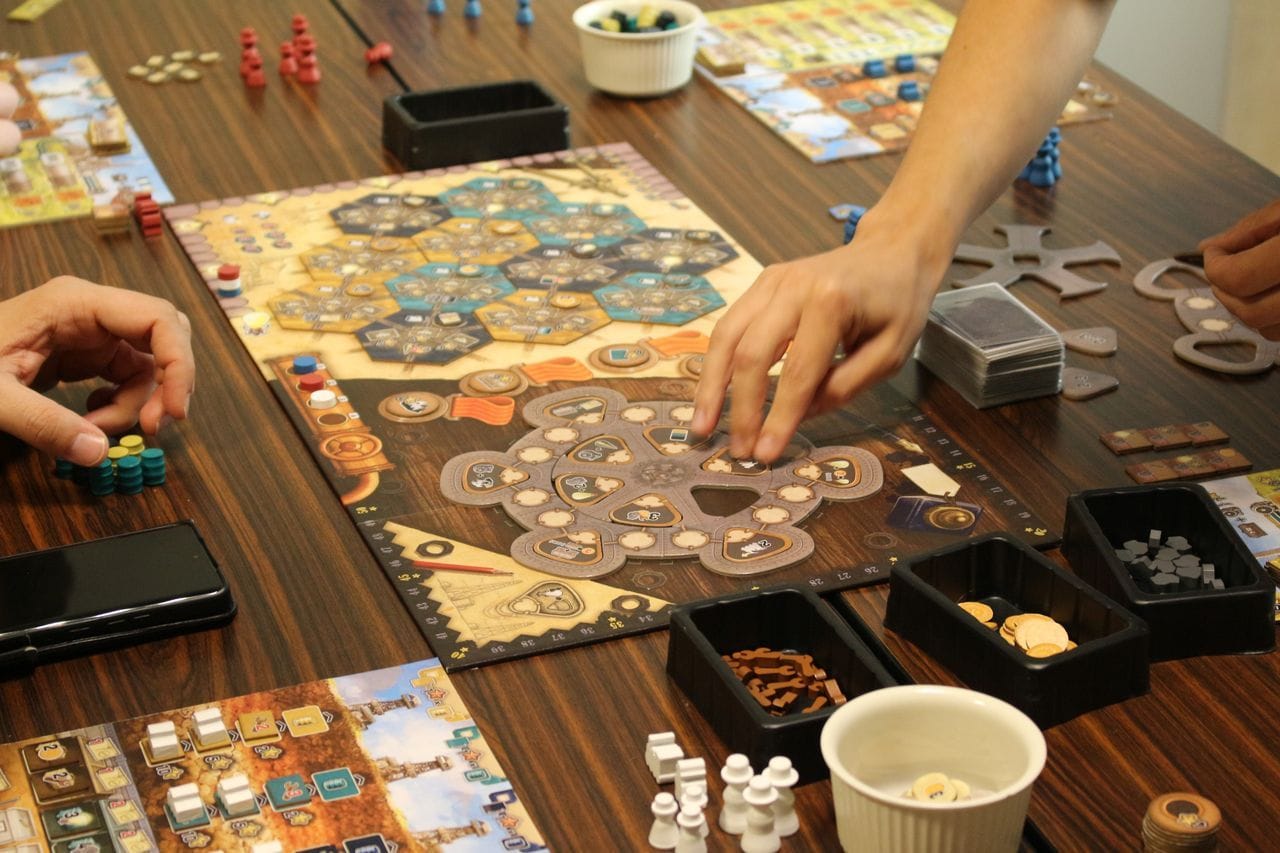 A game board with twelve hexagonal region tiles on it features four goal tokens as well as an action wheel with twelve actions placed in empty slots. Game boards and components for individual players surround it.
