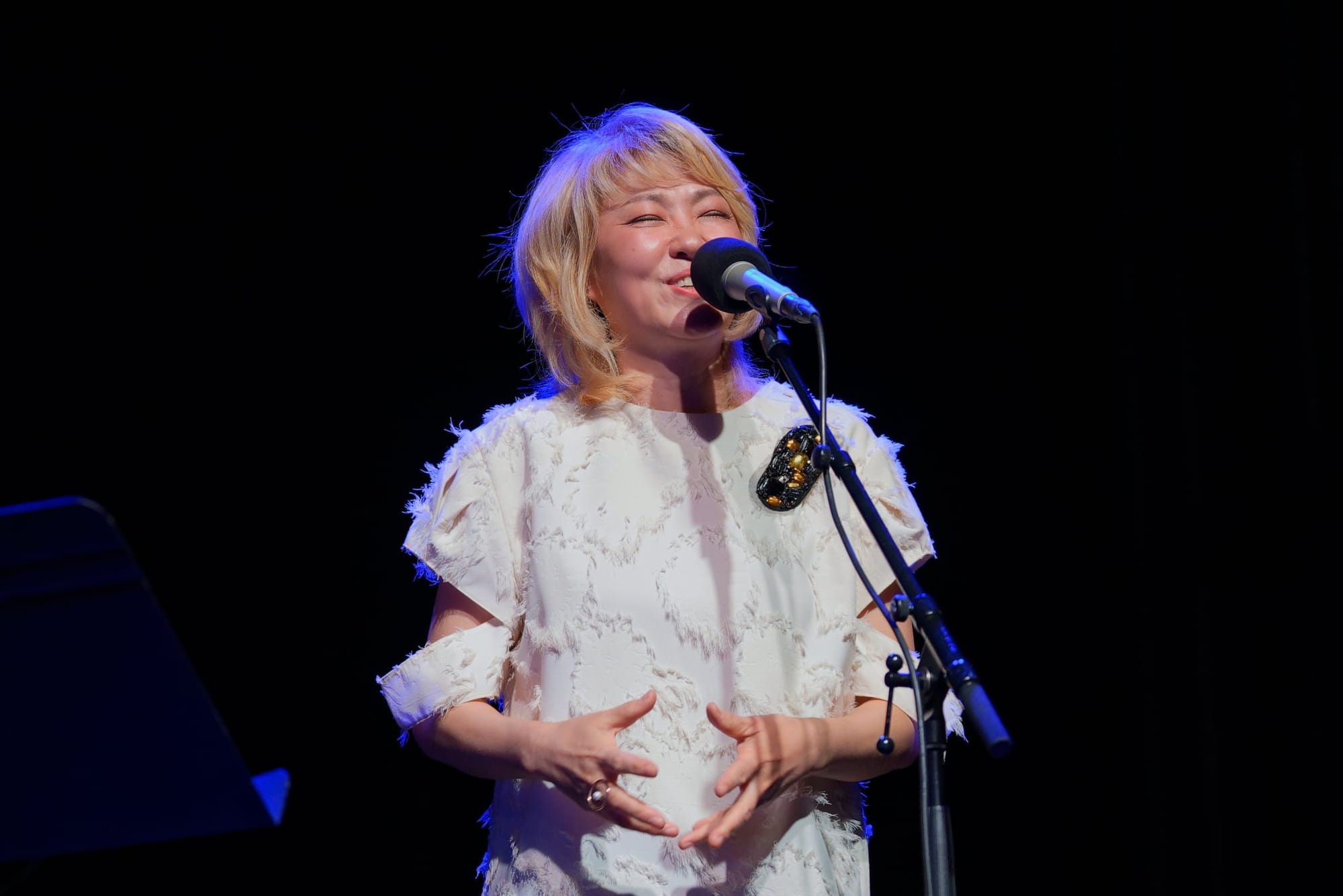 Youn Sun Nah with bleached hair, a white top, and her hands in an expressive gesture, lit up behind a microphone and against a dark background.