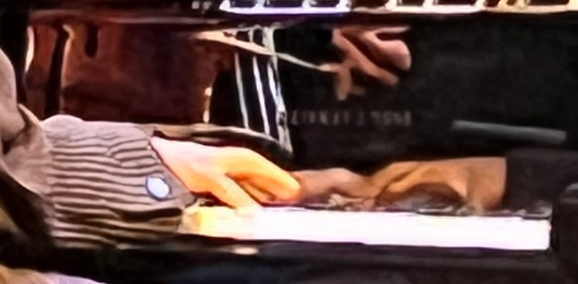 A blurry closeup of hands playing a piano and reflected above the keys, where a distorted of a hand holding a bow being used to play a bass is also visible.
