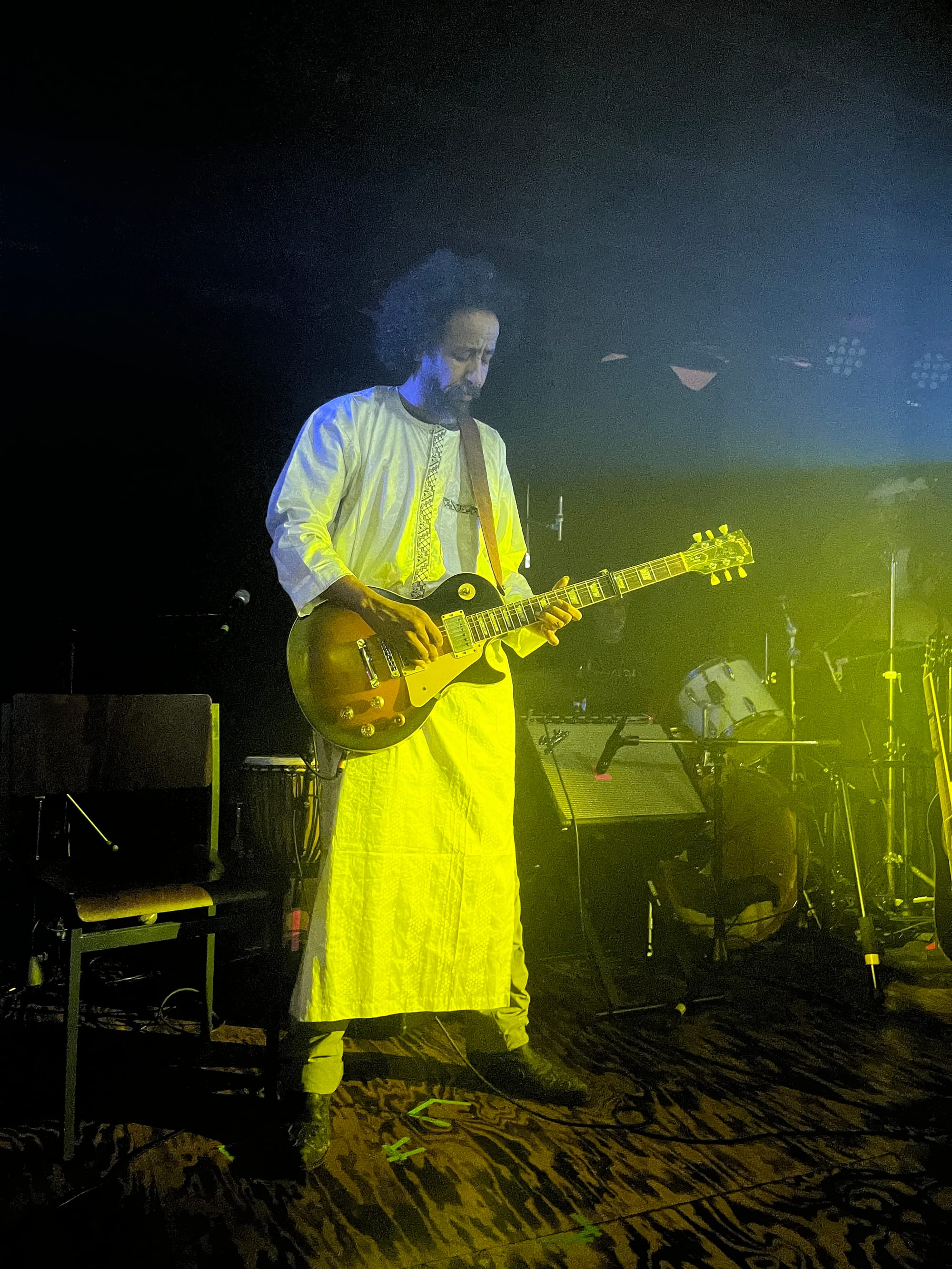 Ousname dressed in a long white robe, playing his electric guitar in shimmering yellow-white light.