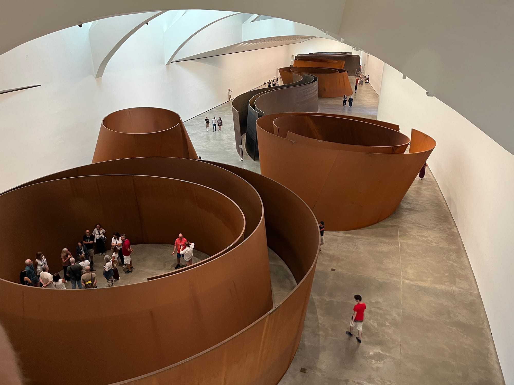 Huge sculptures made of weathered steel curve through the space of a long white museum hall. People are scattered around and inside the sculptures, which you can walk around and through and into.