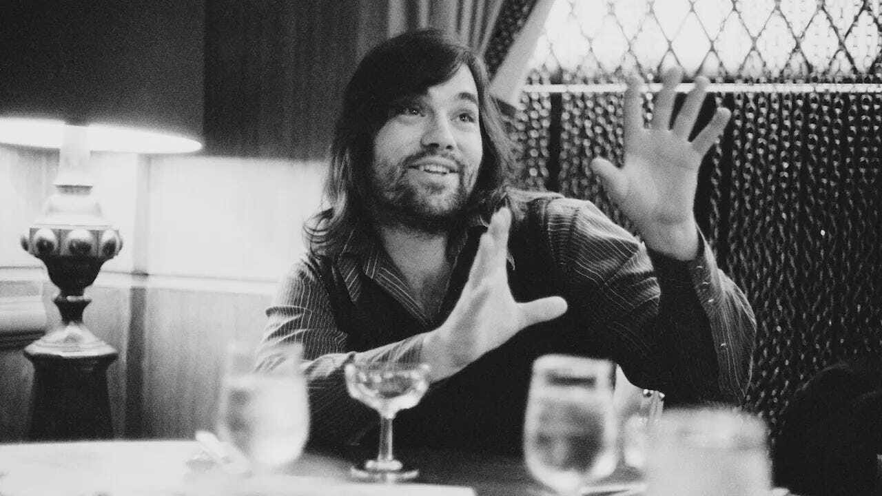 Lowell George sitting at a table holding his hands up in an enthusiastic gesture.
