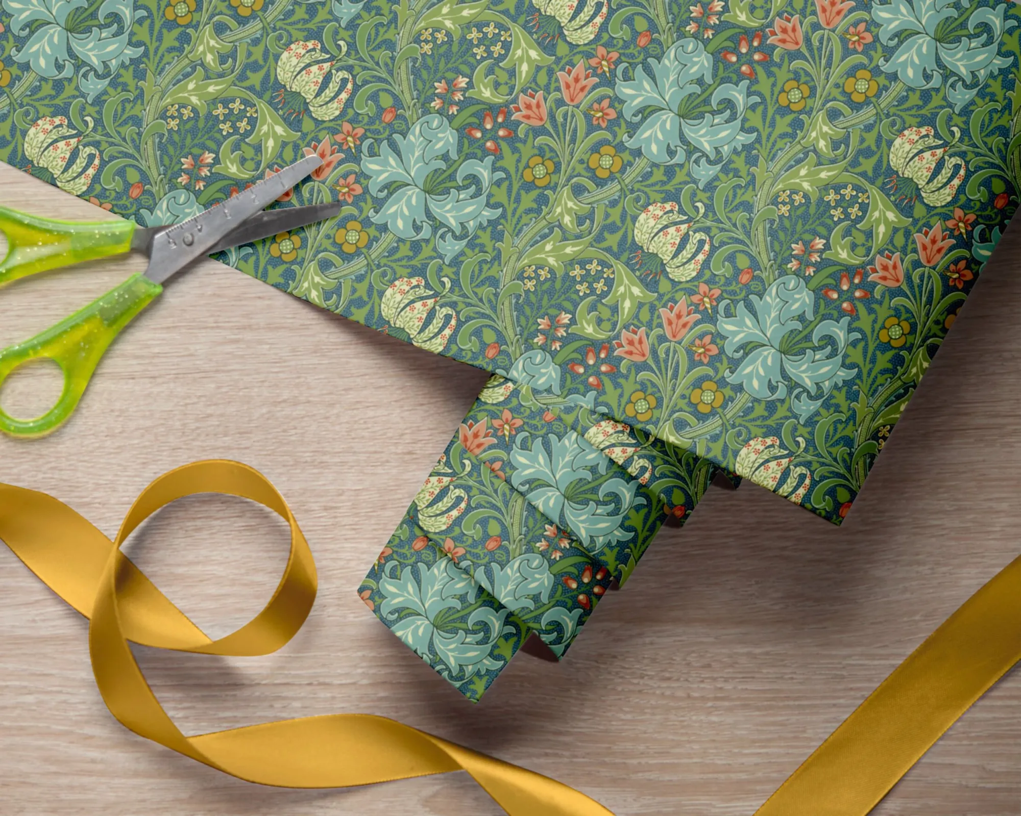Golden Lily wrapping paper folded round a gift, with scissors and gold ribbon on a pale wooden table.