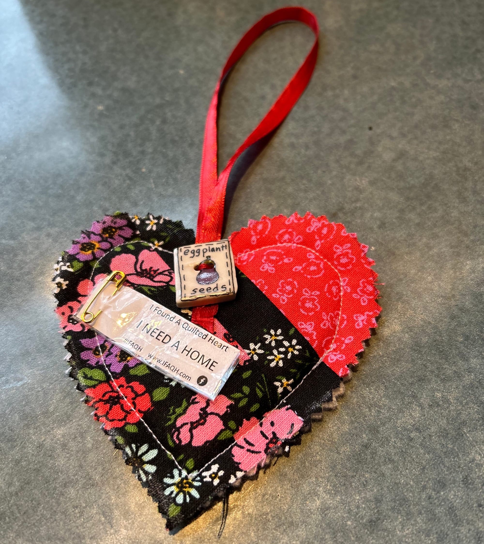A quilted heart found in a local park. It has a button that reads "eggplant seeds" and a tag pinned on that reads "I NEED A HOME." 