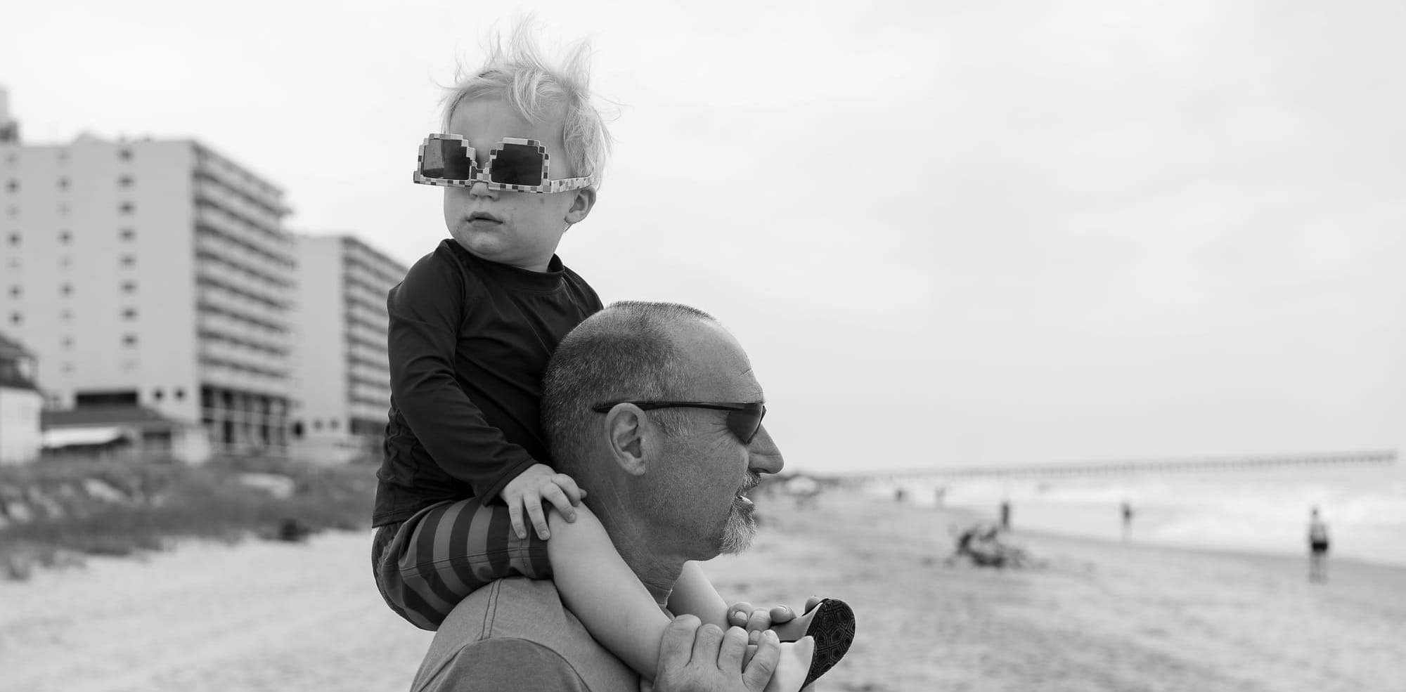 A toddler riding on a man's shoulders gets a view through upside-down sunglasses. They are walking on a beach. 