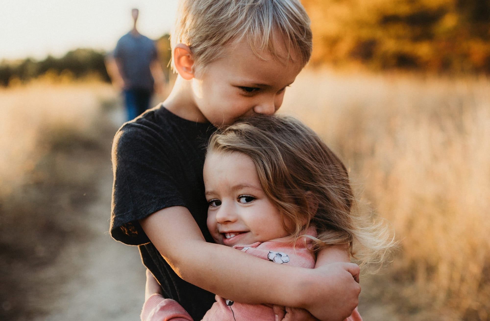 A young boy hugs a younger girl, dried grassy field and an adult in the background.