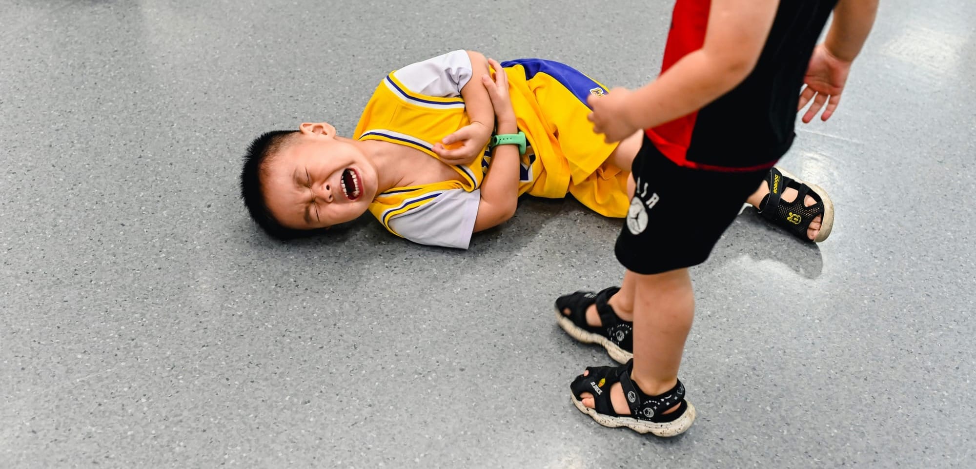 Young boy in sports uniform on the floor crying. Another child stands by his side.