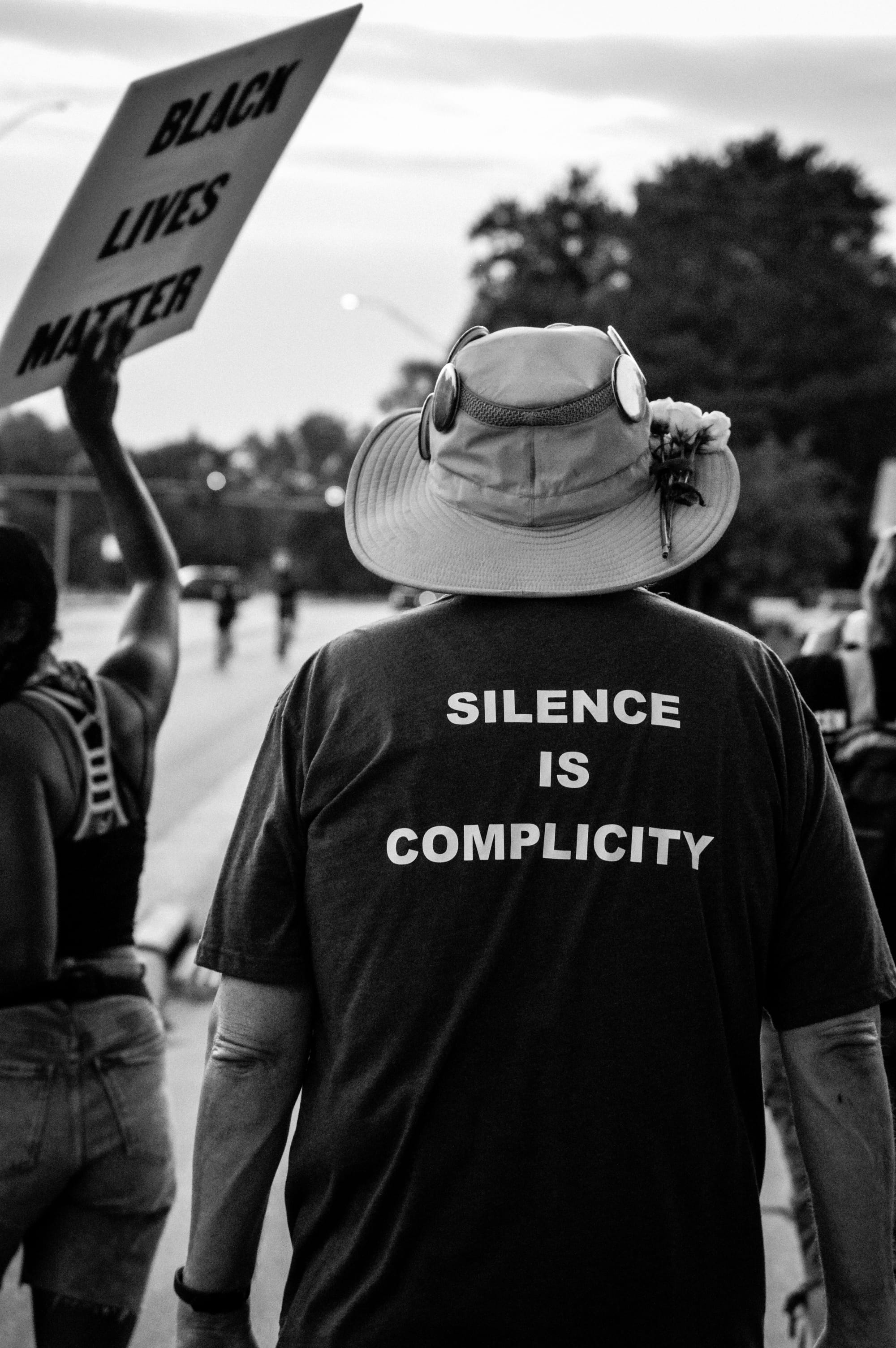 One person holds up a poster reading "Black Lives Matter," another's T-shirt reads "Silence is Complicity." Both are facing away from the camera.