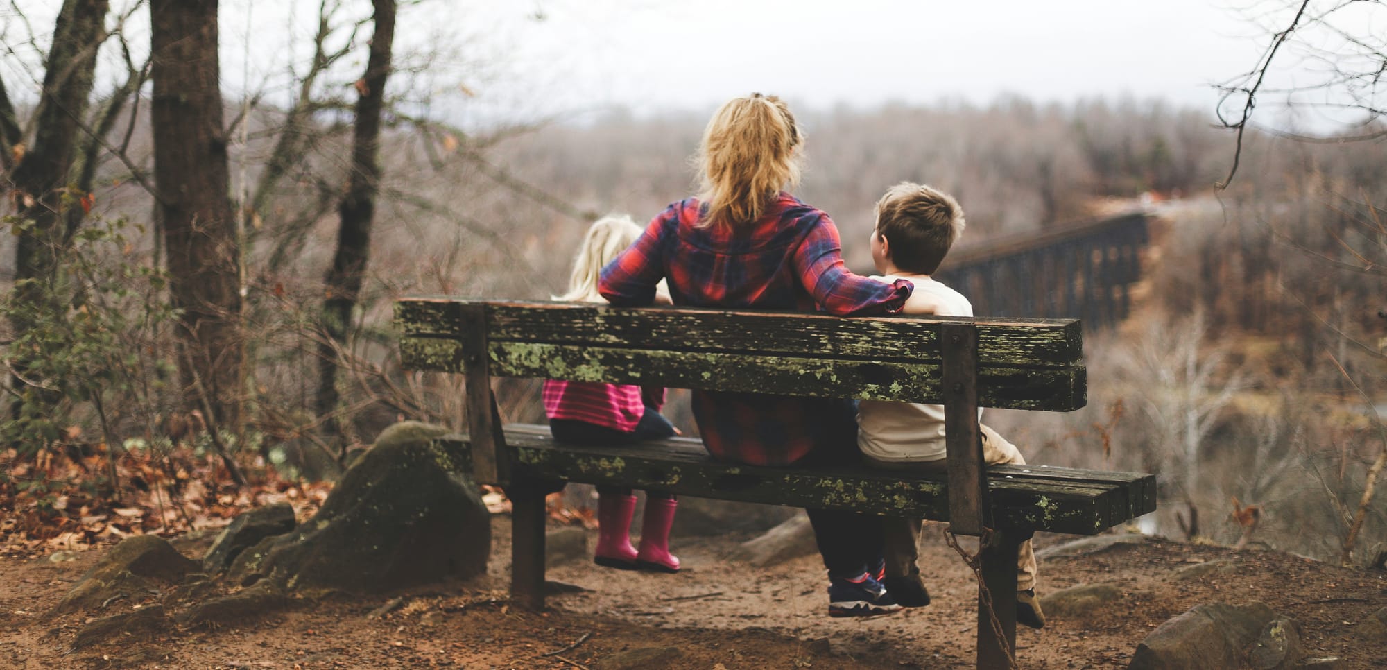 A woman sits between two children on a bench in a hilly, woodsy late fall setting. 