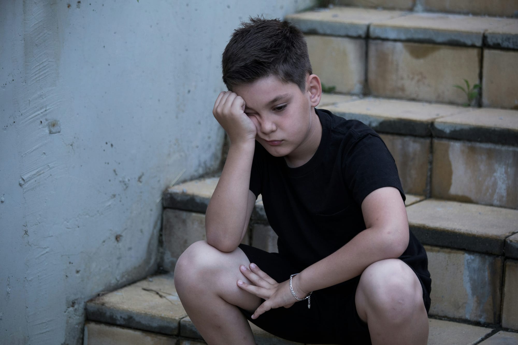 A boy sits on steps outdoors, head in his hand, looking glum.