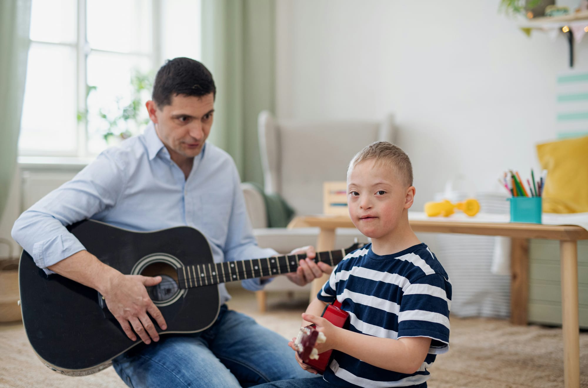 A boy, perhaps with Down syndrome, plays his guitar with an adult who also has a guitar.