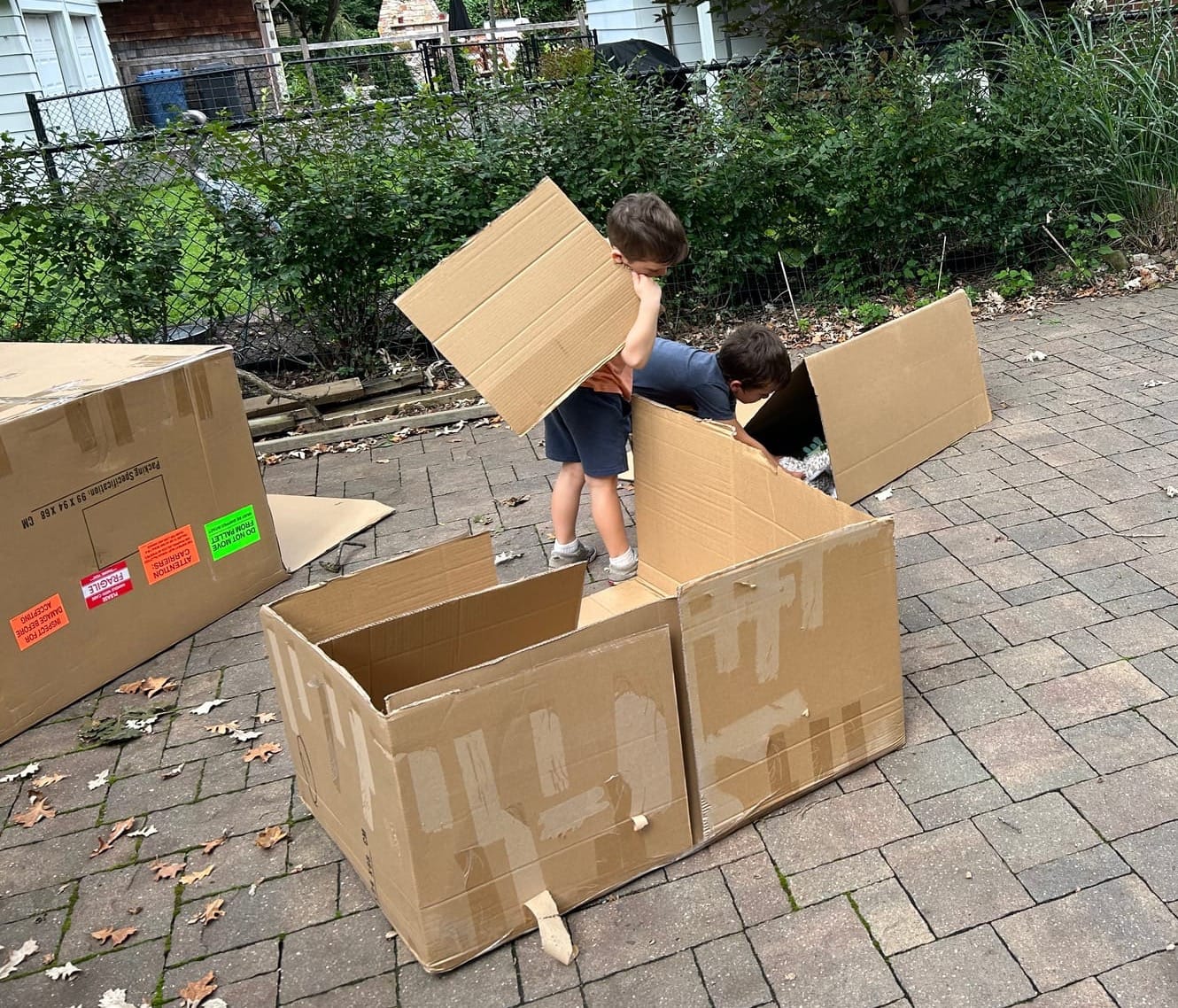 Two boys make homes for their stuffies out of large cardboard boxes.