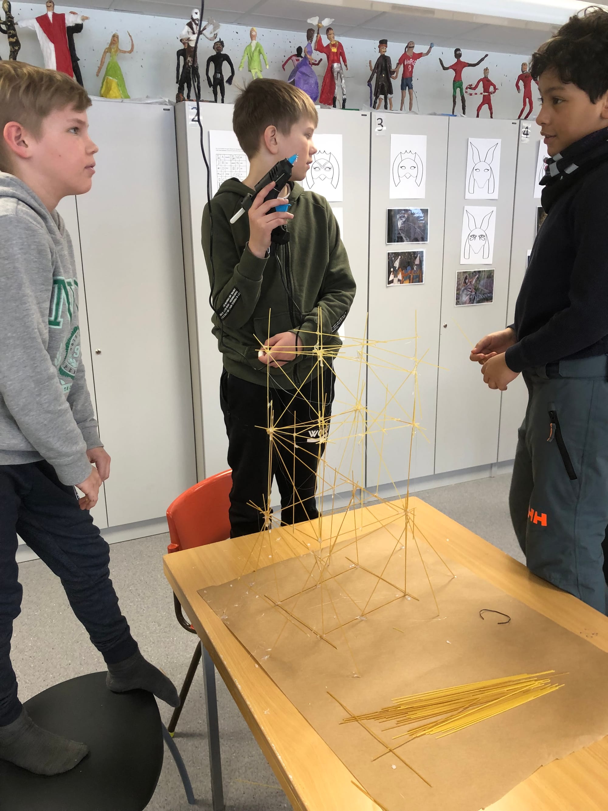 Three middle-school boys work on building a spaghetti tower. So tall they are standing on chairs; one holds a glue gun. 