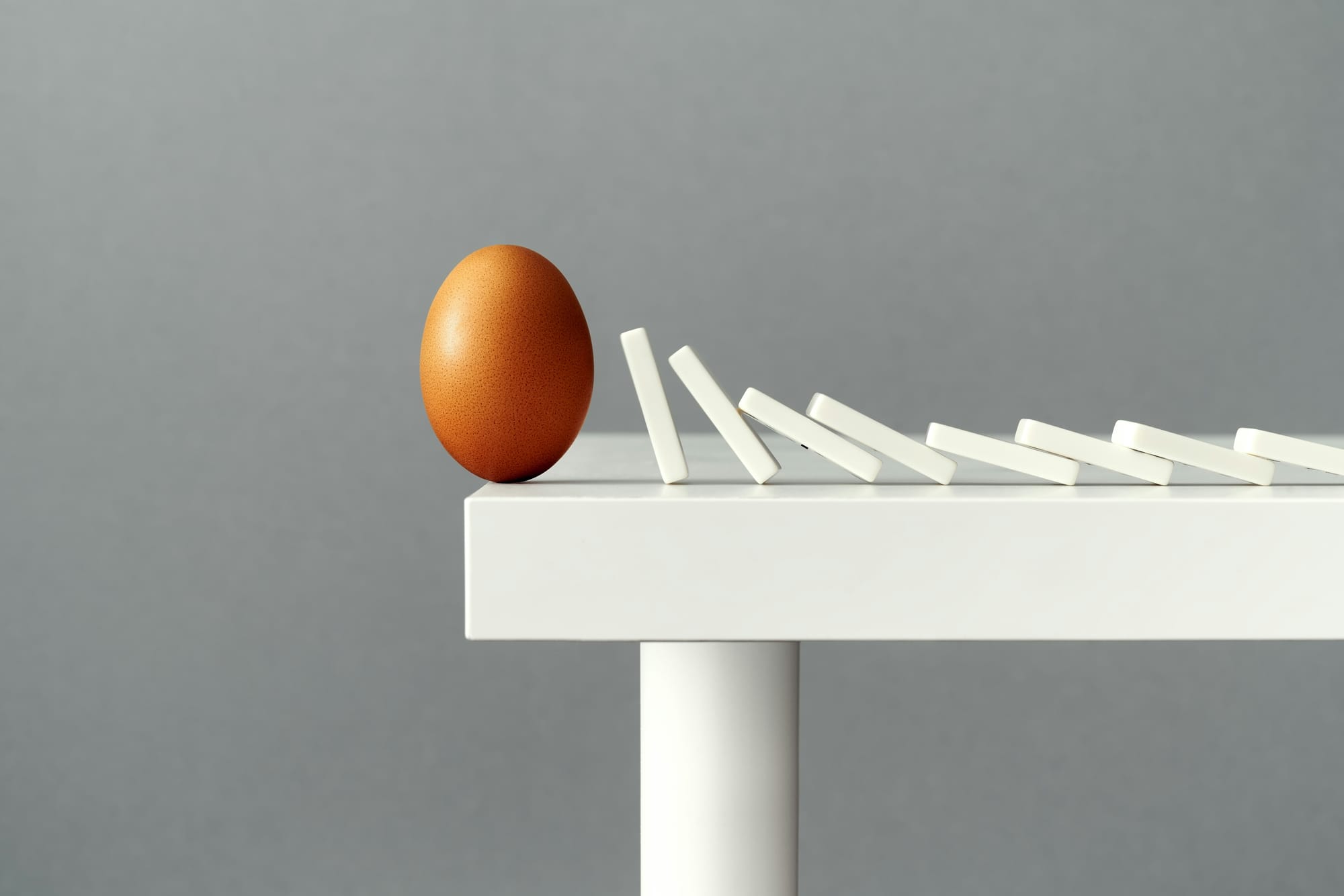 An upright brown egg on a white table, about to be toppled to the floor by falling dominos. 