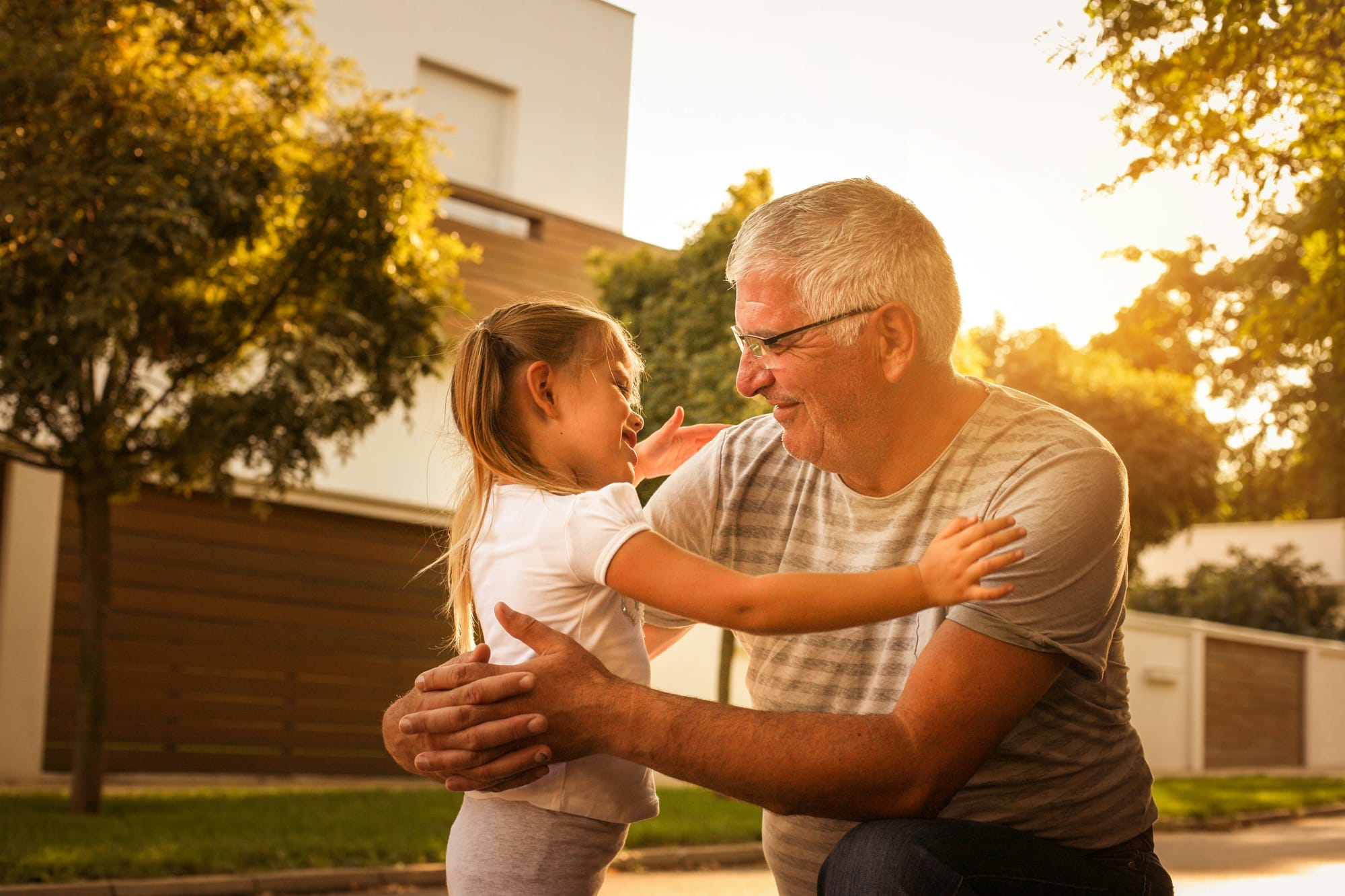 A grandfather and grandchild share a hug in an outdoor, sunny setting.