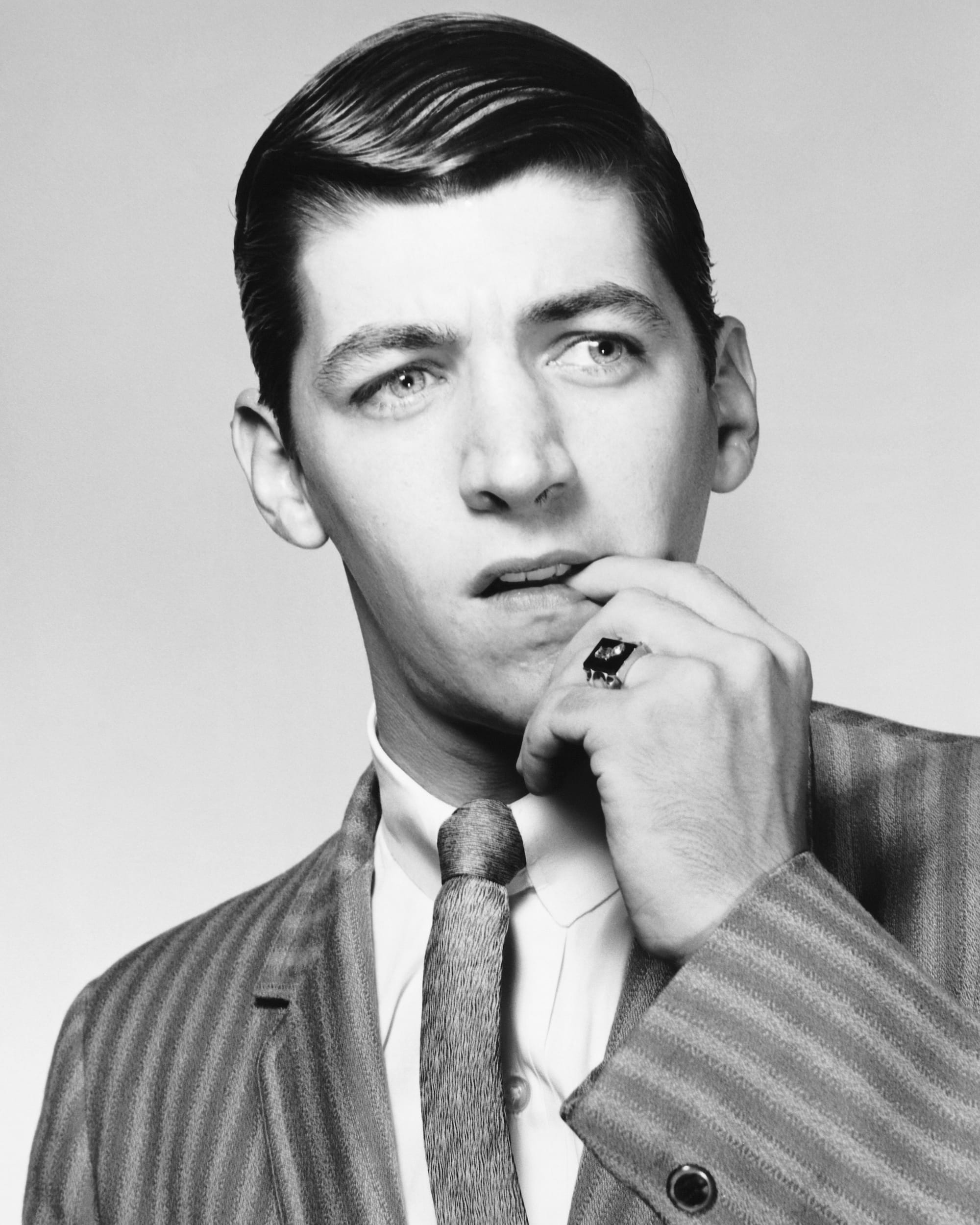 Black and white photo of man in a suit, finger in his mouth, appearing hesitant. 