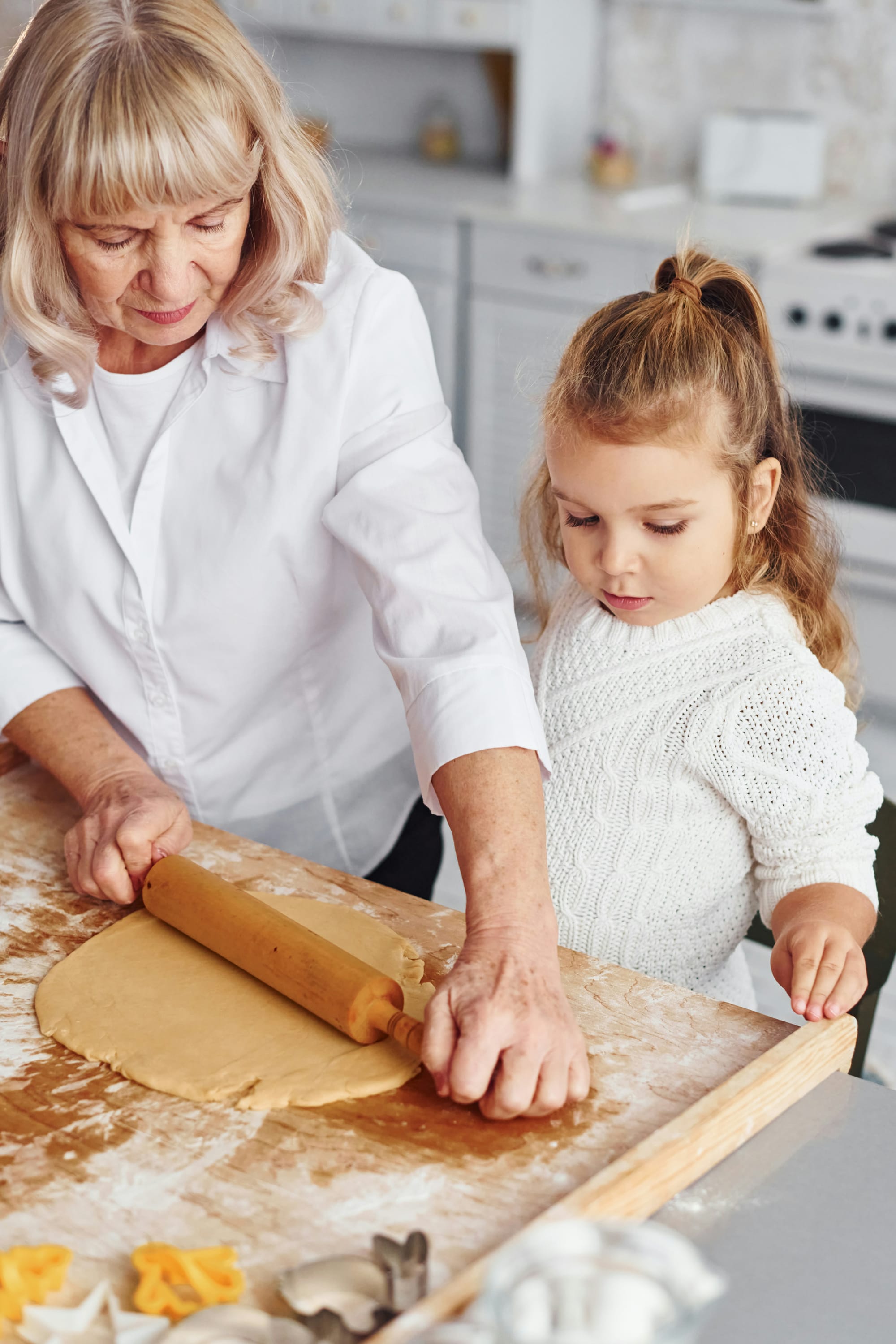 A grandmother and granddaughter rolling out cookie dough on a wooden cutting board.