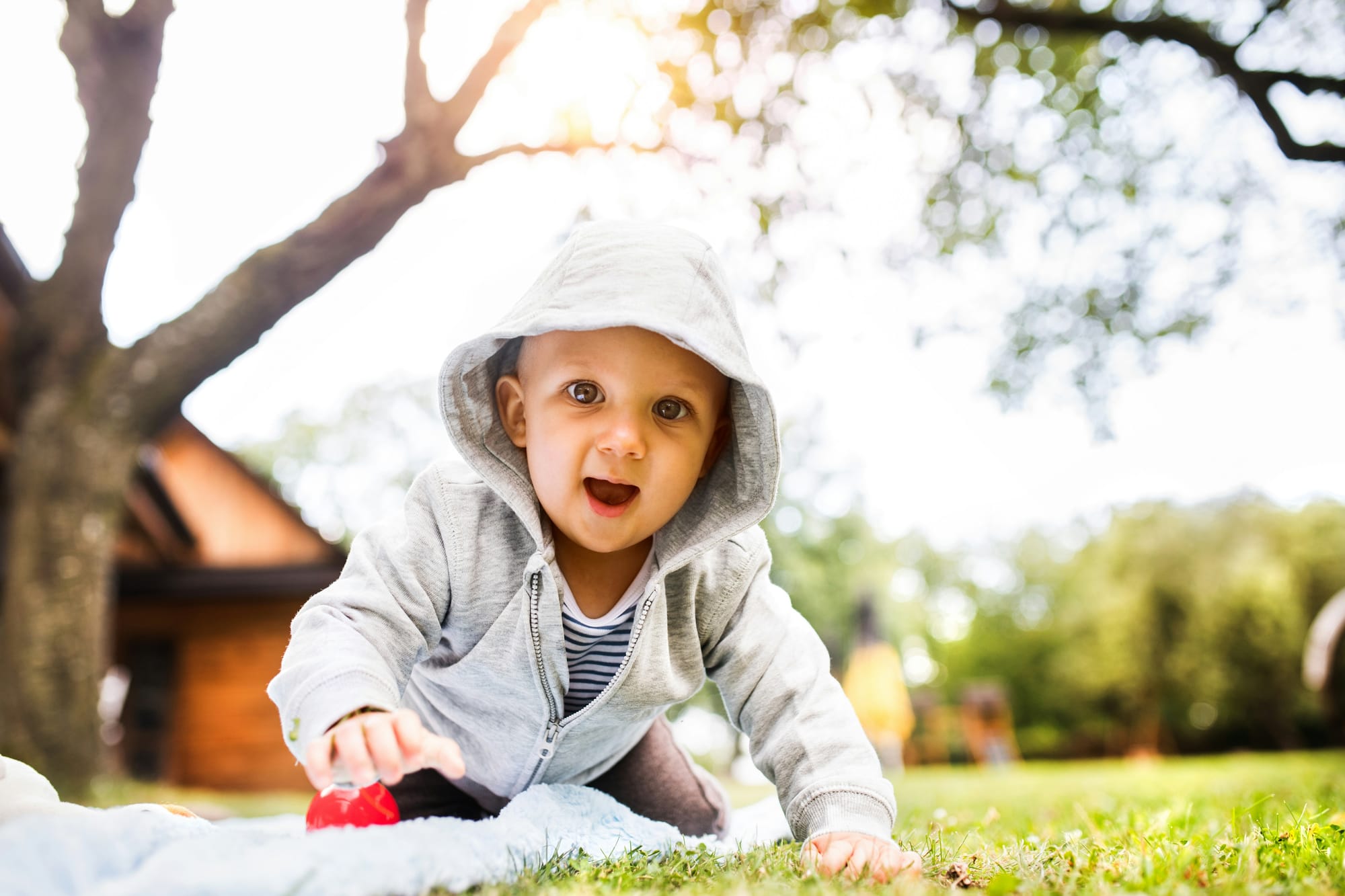 A baby wearing a hoodie smiles, playing with toys outside on a blanket on a lawn.