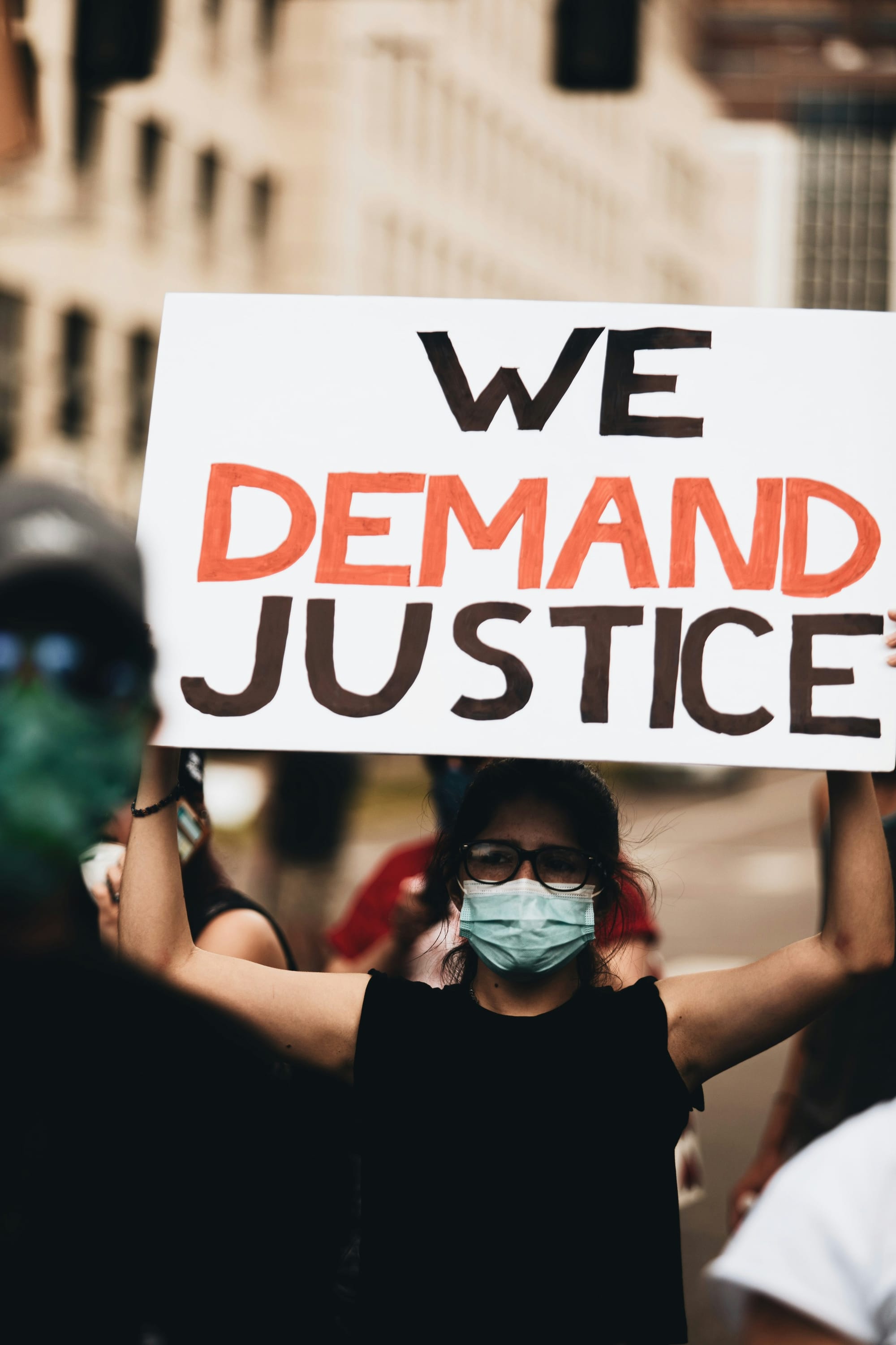  A woman in a crowd, wearing a black t-shirt and medical mask, holding up a sign that reads, "We demand justice."