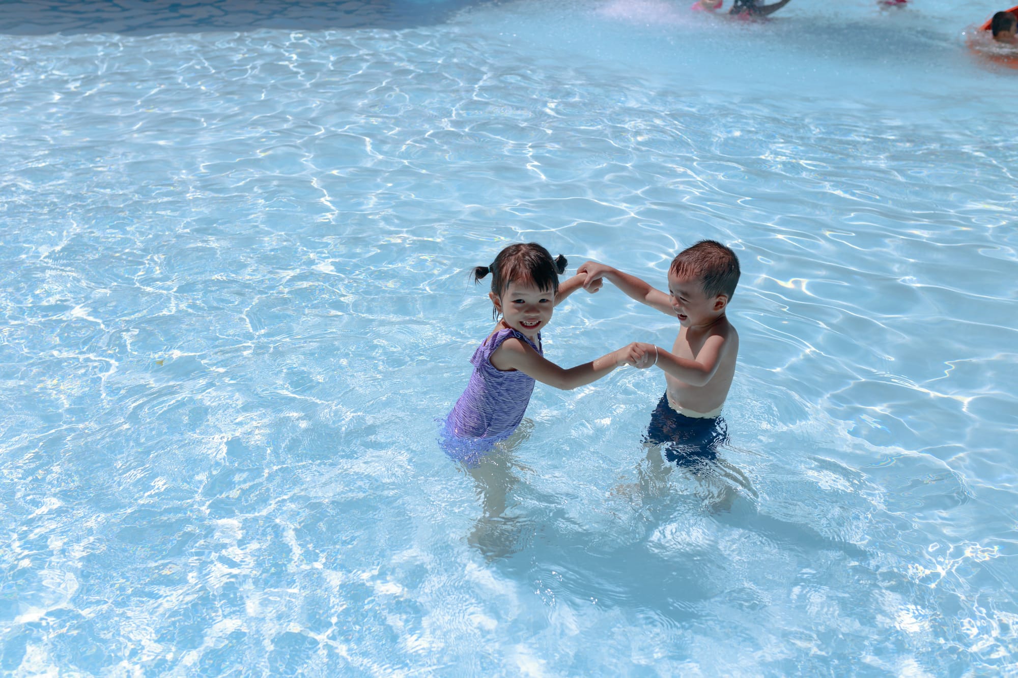 Two smiling preschoolers hold hands in a sunny wading pool.