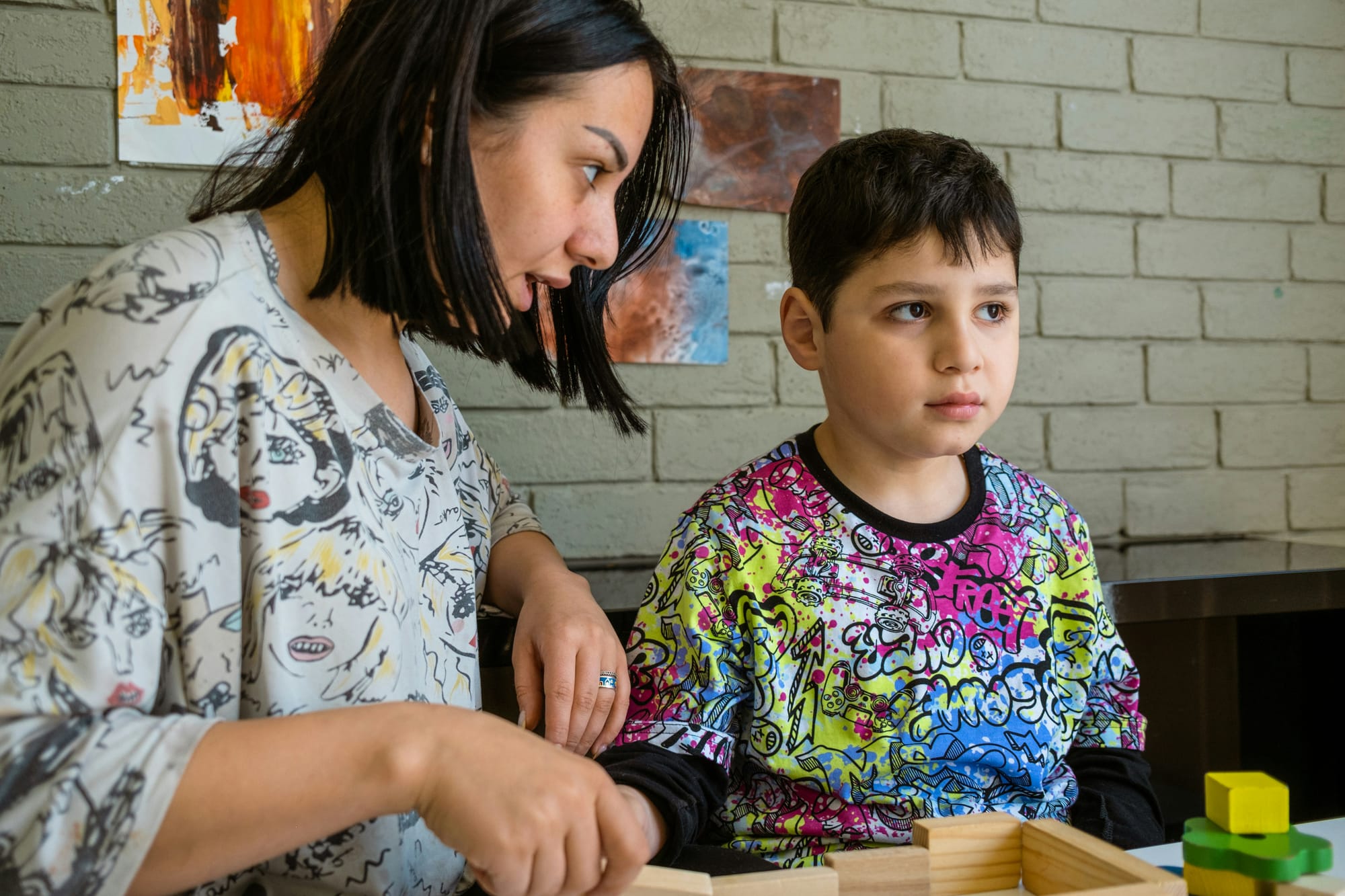 A grade-school age boy sits with a woman, who is speaking to him. They are building with blocks.