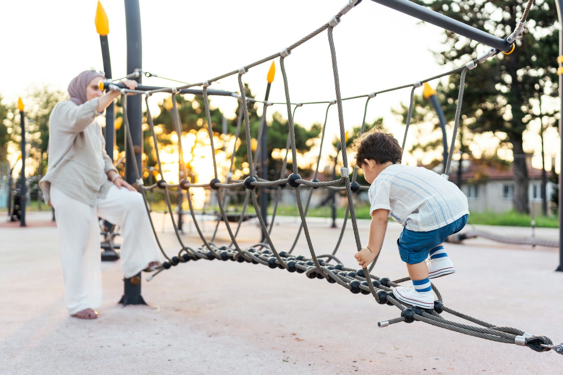 A preschool-age boy starts through a net climbing toy at a park. A woman stands waiting at the other end.