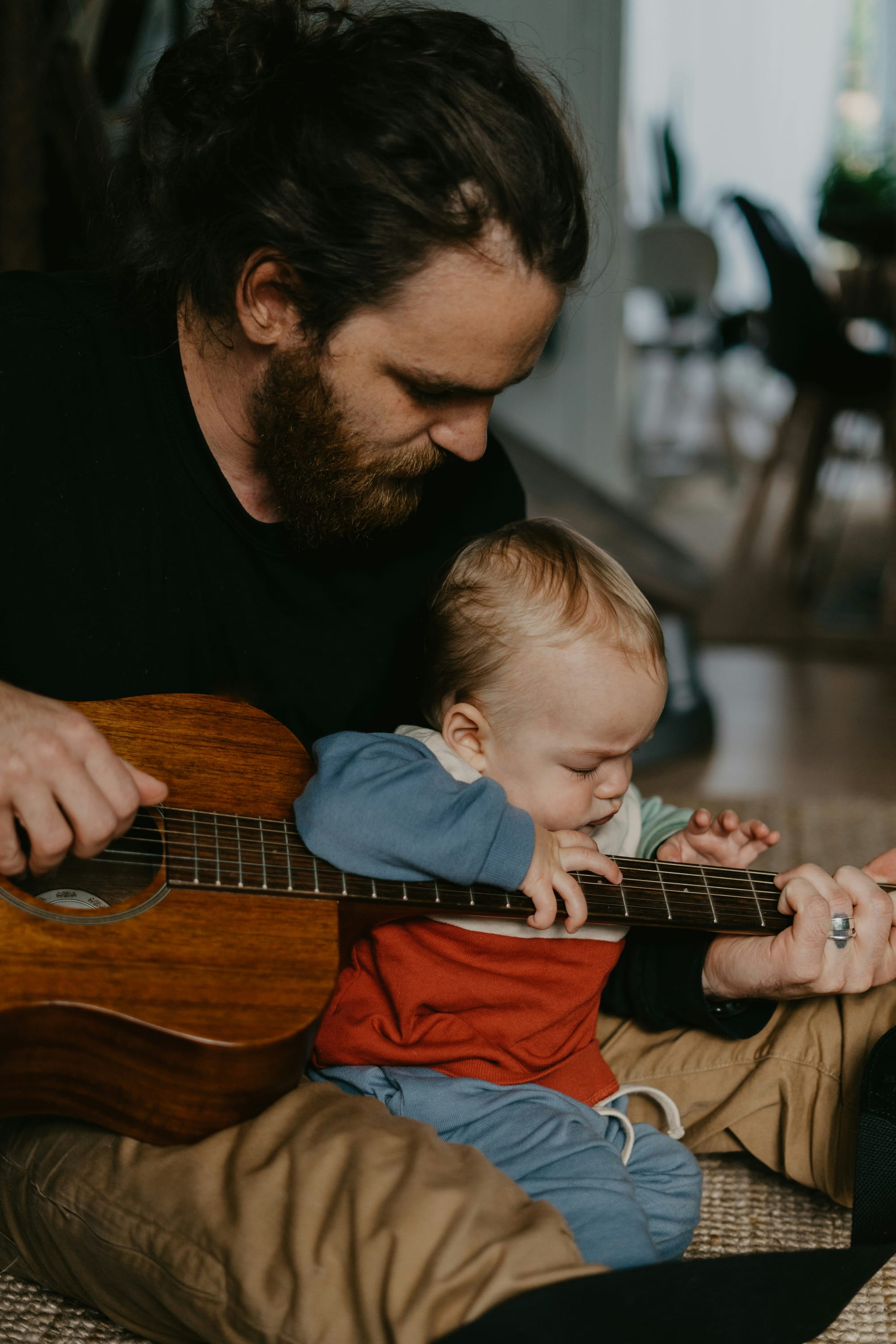 A baby tries a guitar siting in the lap of man who is holding guitar and baby.