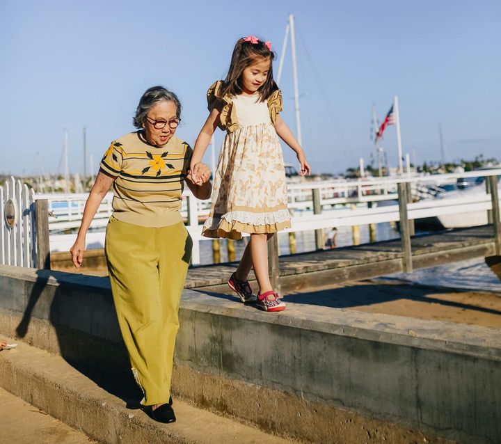 A grandmother helps her grandchild balance on a cement barrier near docks and a beach.